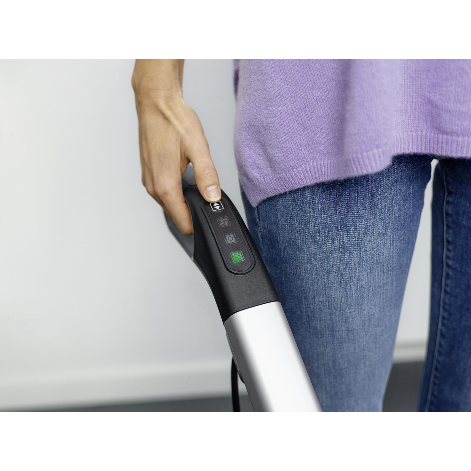 De hand van een persoon drukt op een knop van een stofzuiger. De persoon draagt een paarse trui en jeans.