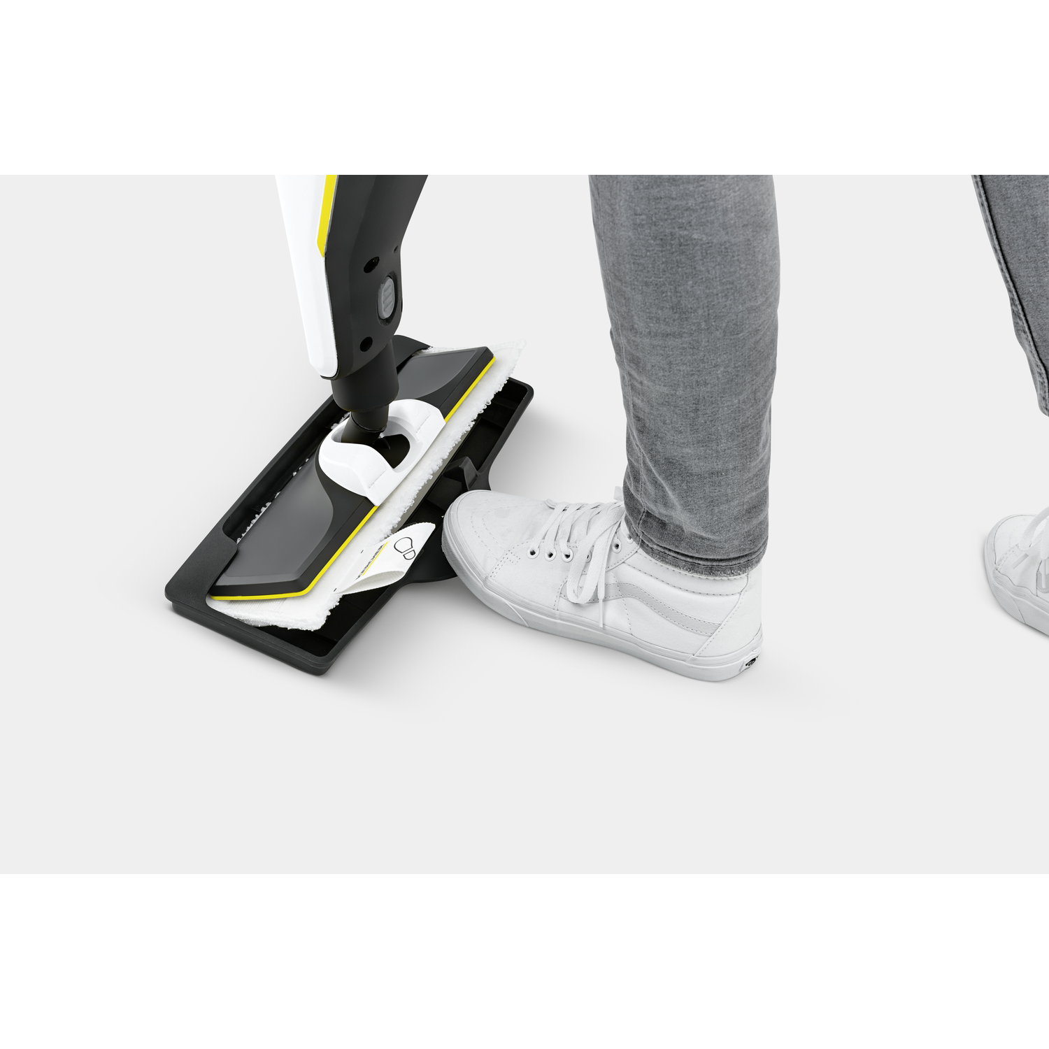 Een persoon gebruikt een zwart-gele vloerreiniger. Ze draagt een grijze jeans en witte sneakers. De achtergrond is wit.