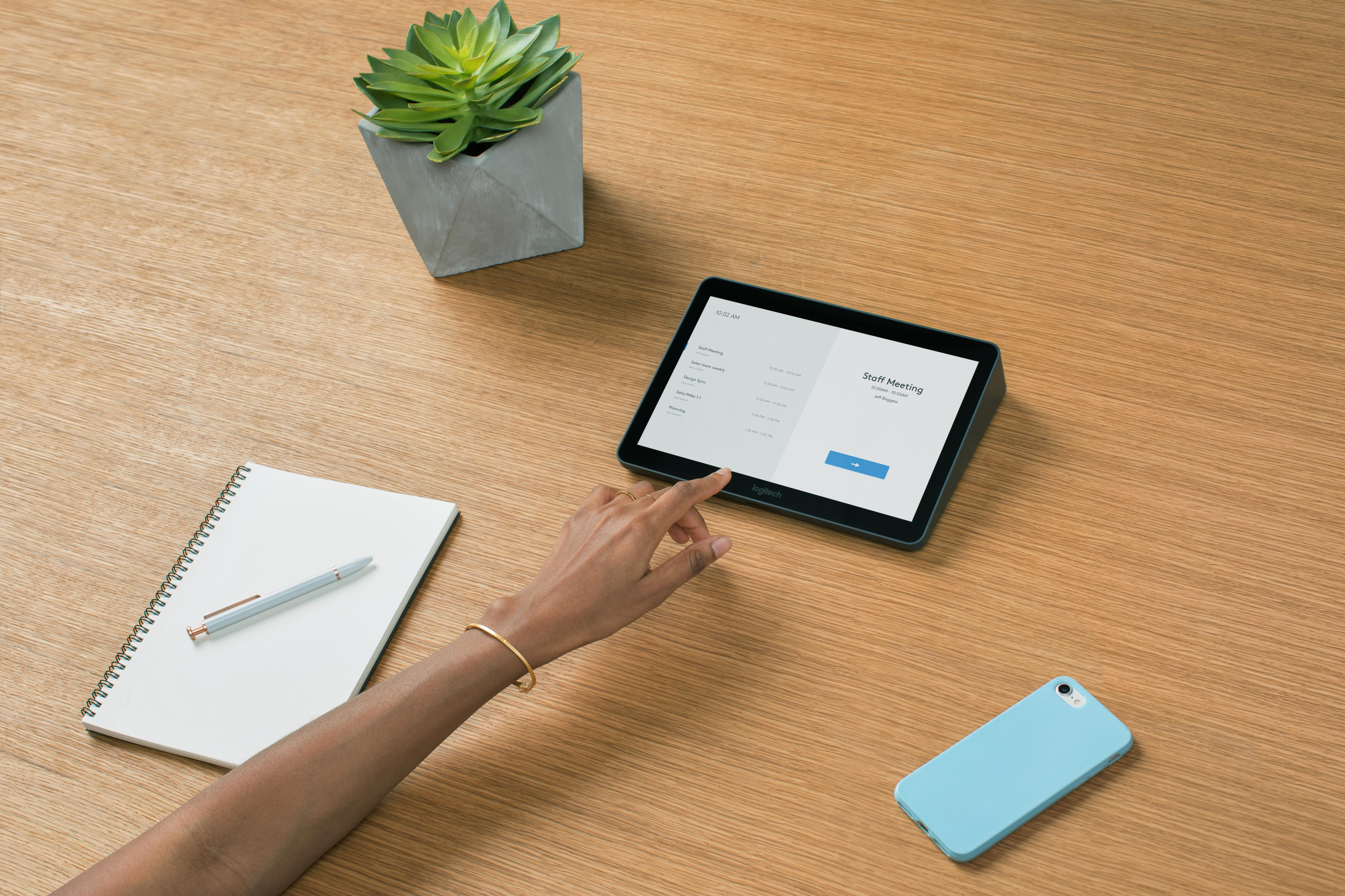 Eine Hand berührt ein Tablet mit 'Staff Meeting' auf dem Bildschirm. Ein Notizblock, Stift, Telefon und Pflanze liegen auf einem Holztisch.