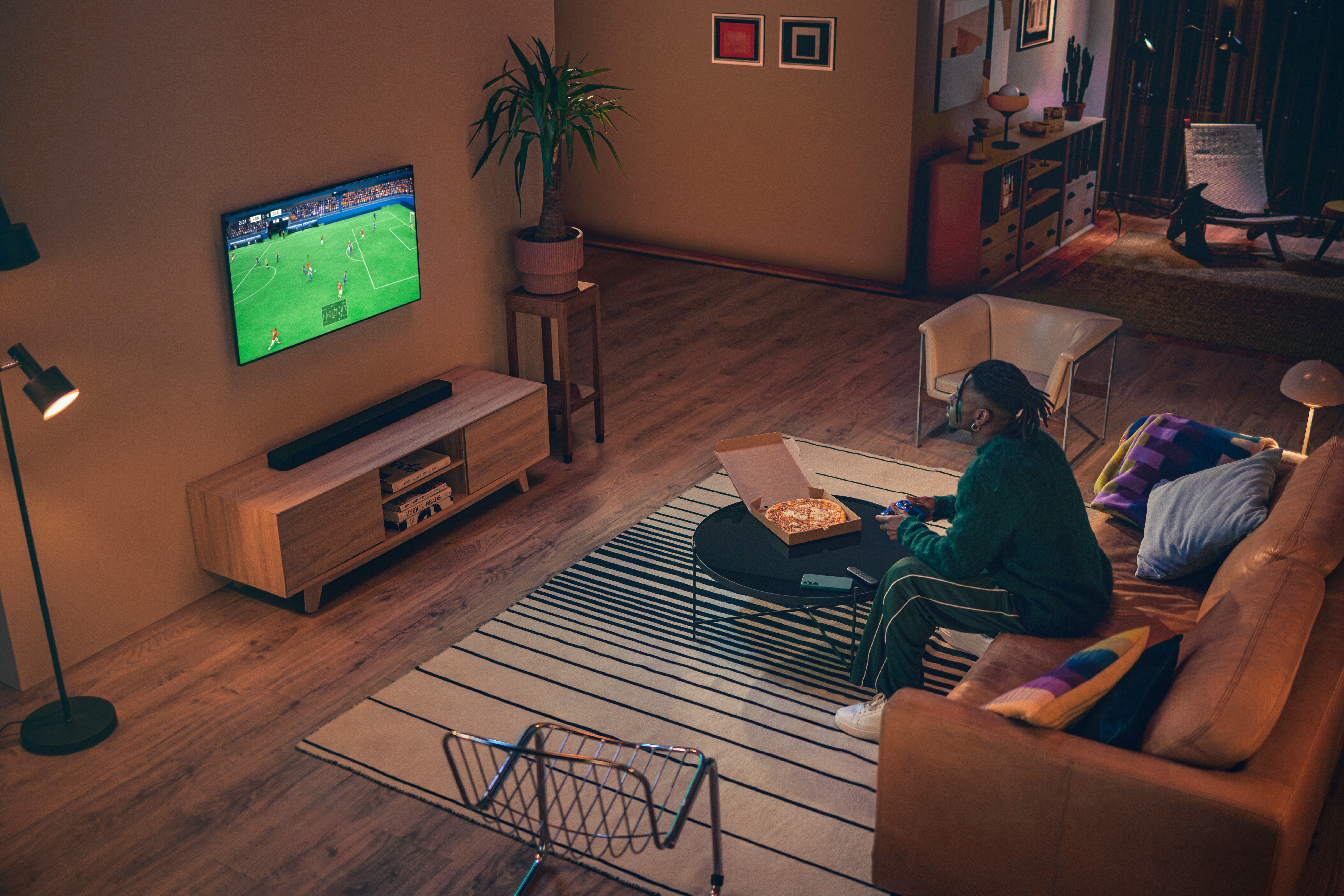 Eine Person sitzt auf einem Sofa und spielt ein Videospiel. Eine Pizza und ein Fernseher mit Fußball sind sichtbar. Der Raum hat Holzböden und einen Teppich.