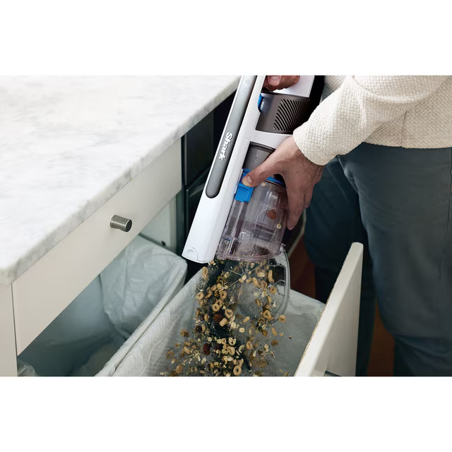 Una persona vacía una aspiradora Shark en un cubo de basura. Los escombros caen de la aspiradora. La escena es en una cocina blanca.
