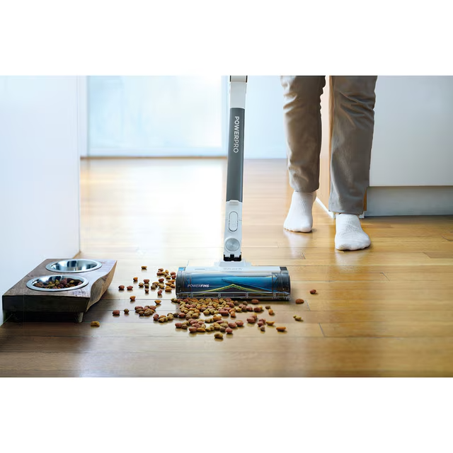Una persona aspira comida para perros esparcida en un piso de madera. Se está utilizando una aspiradora. Un cuenco para perros está a la izquierda.