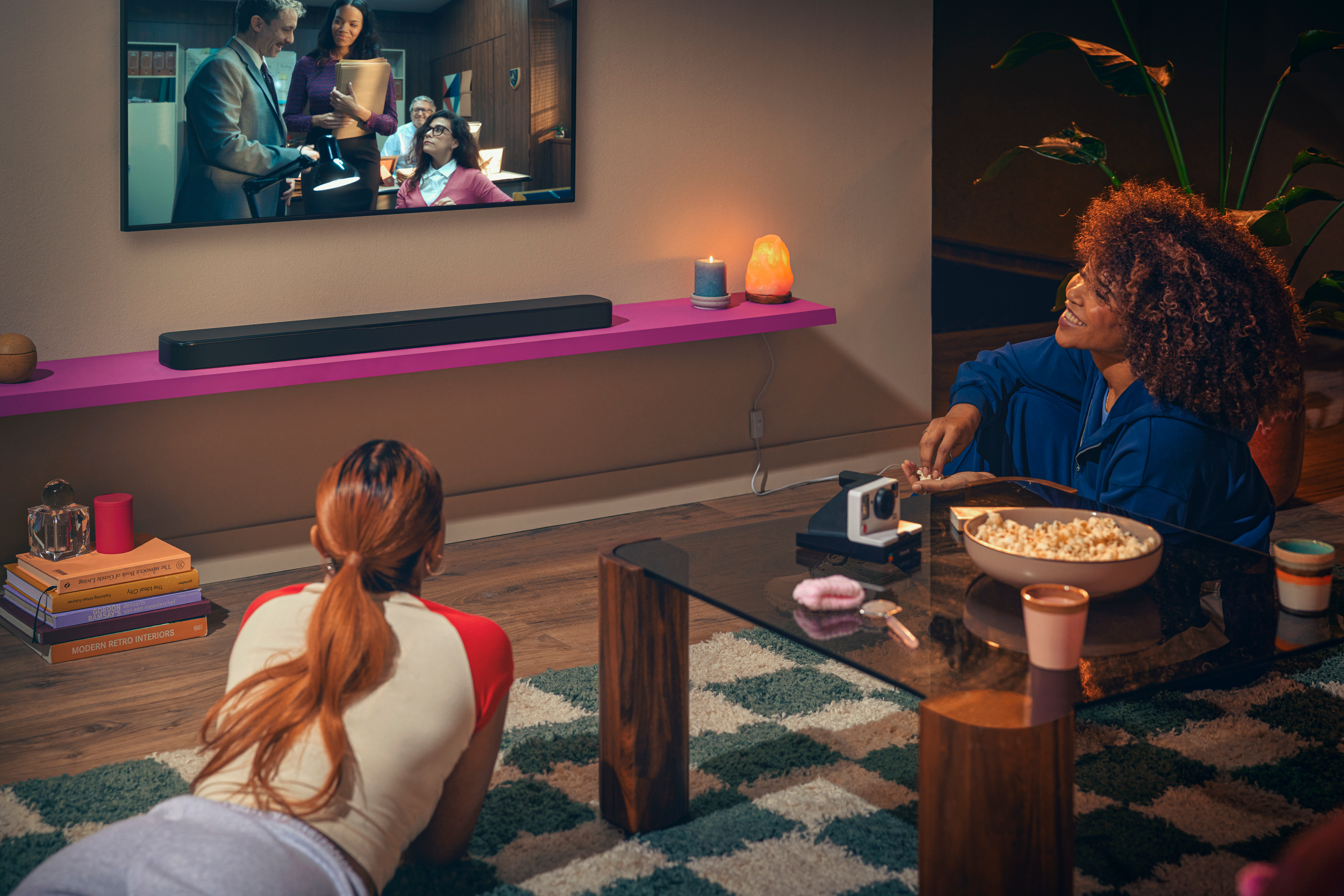 Zwei Frauen entspannen sich. Eine liegt, die andere sitzt mit Popcorn. Im Hintergrund läuft ein Fernseher.