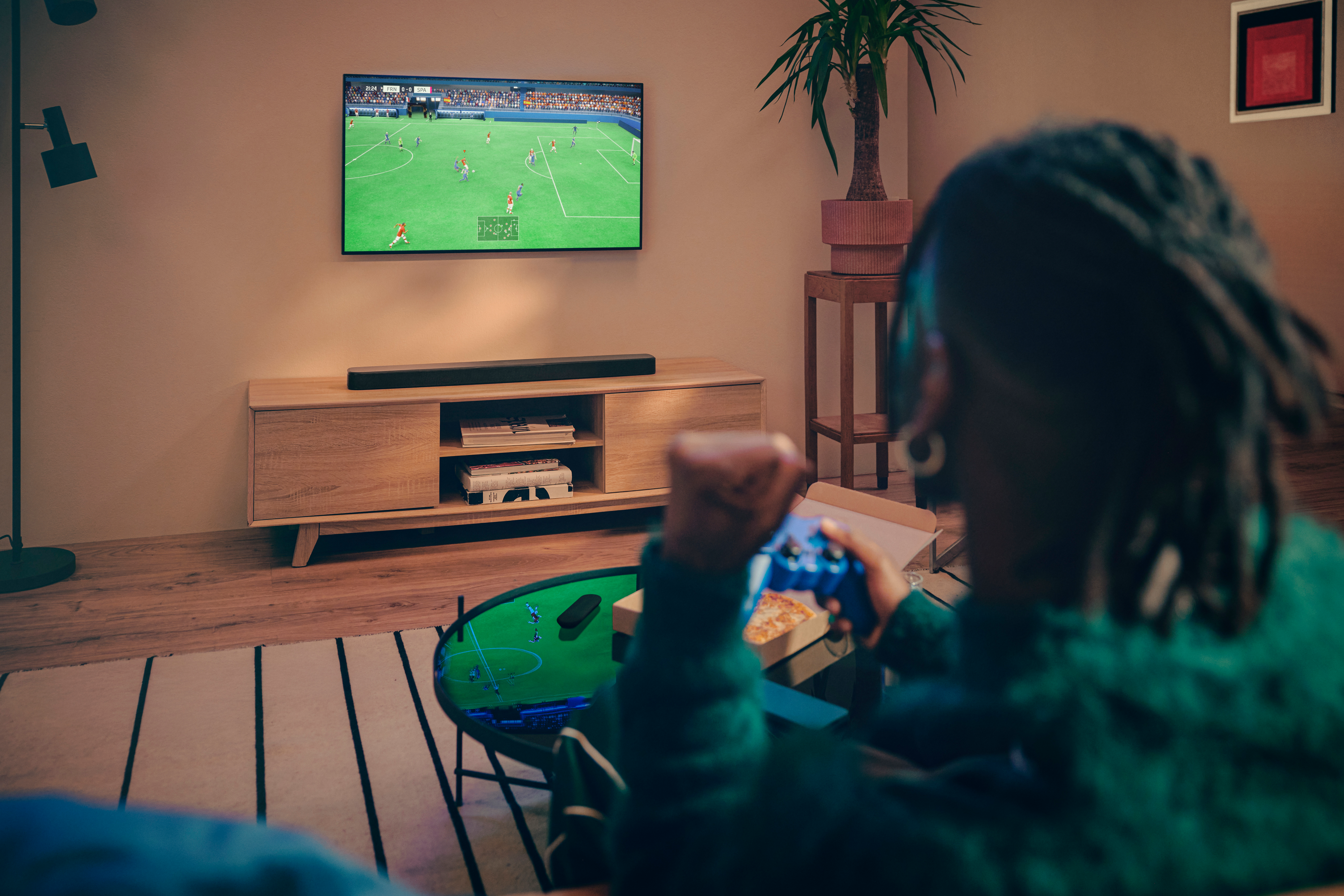 Eine Person spielt ein Videospiel und sieht sich ein Fußballspiel im Fernsehen an. Pizza und ein Controller befinden sich im Vordergrund.