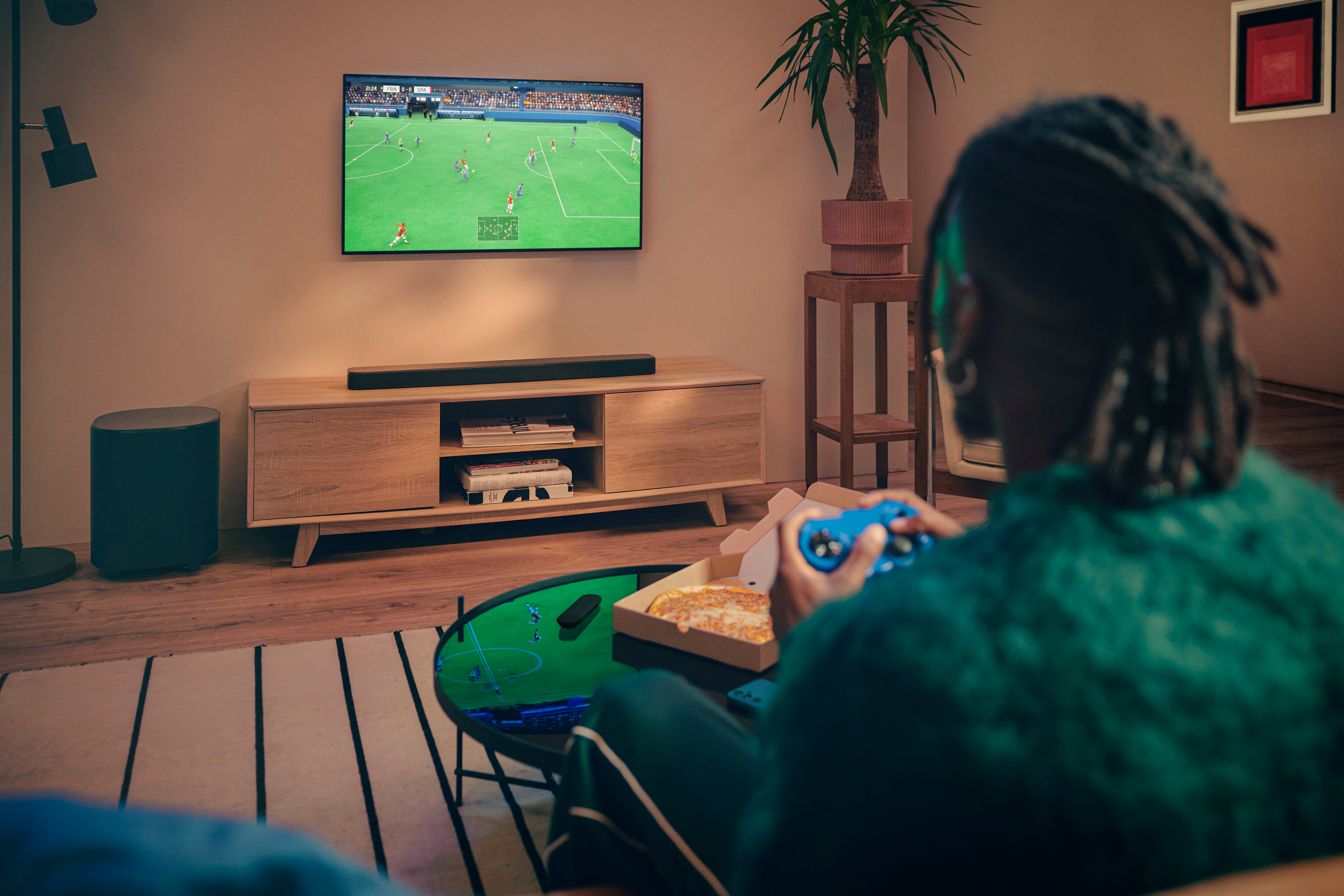 Una persona gioca a un videogioco di calcio in TV, tenendo un controller blu. Pizza su un tavolo di fronte a loro.