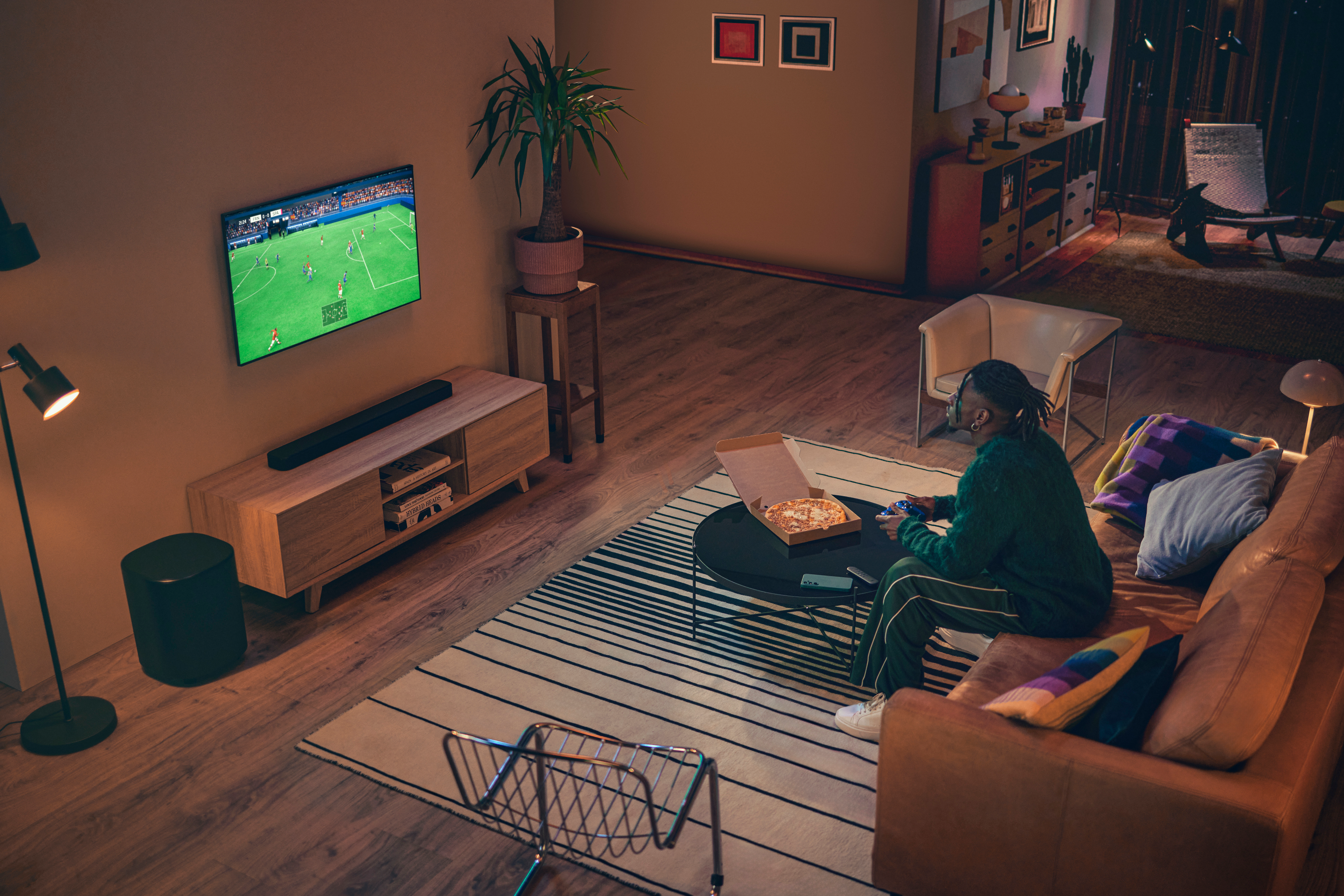Una persona seduta su un divano gioca ai videogiochi. Una pizza e una TV sono visibili. La stanza ha pavimenti in legno e un tappeto a strisce.