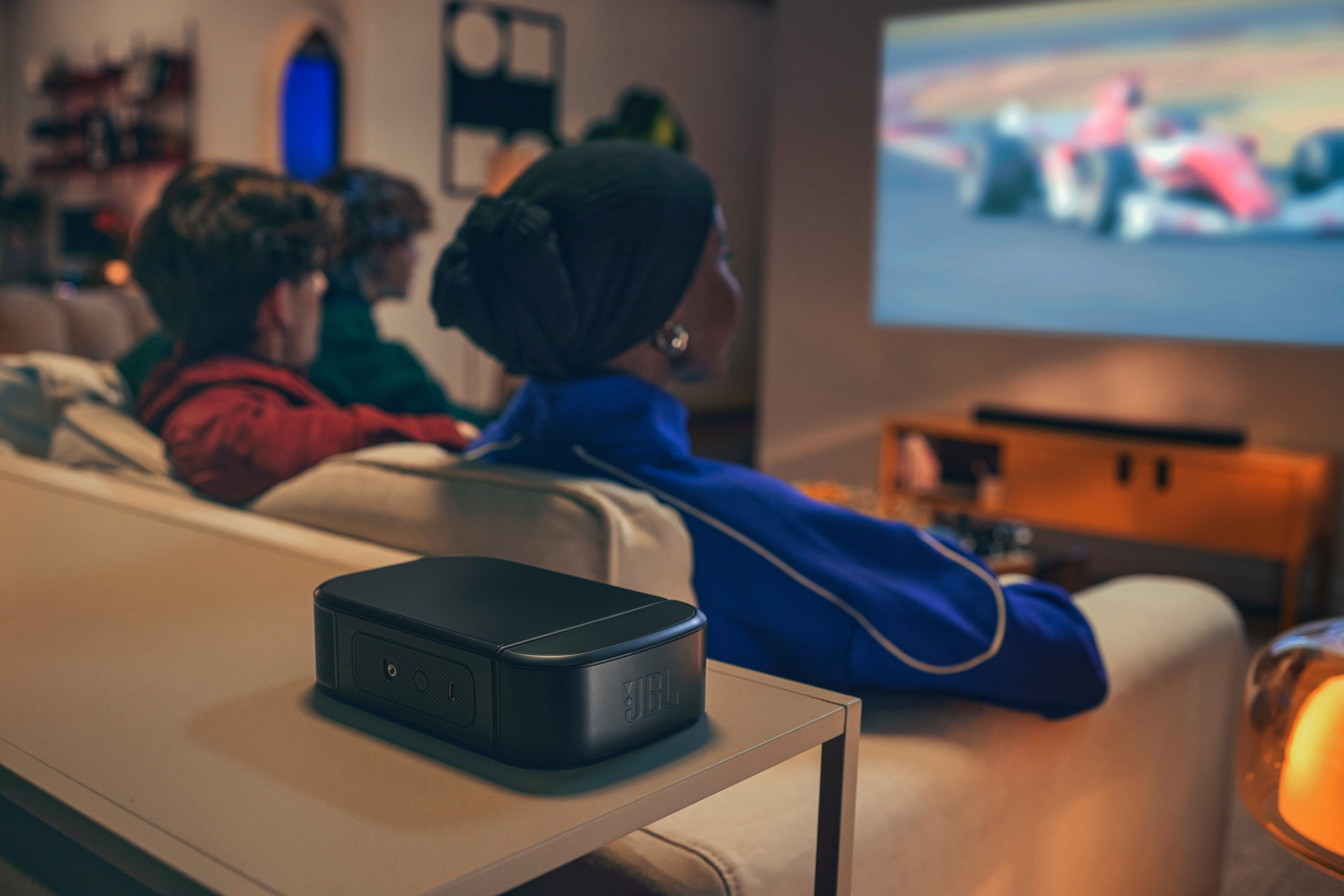 Menschen schauen ein Rennen auf einer Projektionsleinwand. Ein schwarzer JBL-Lautsprecher steht auf einem Tisch. Zuschauer sitzen auf einem beigefarbenen Sofa.