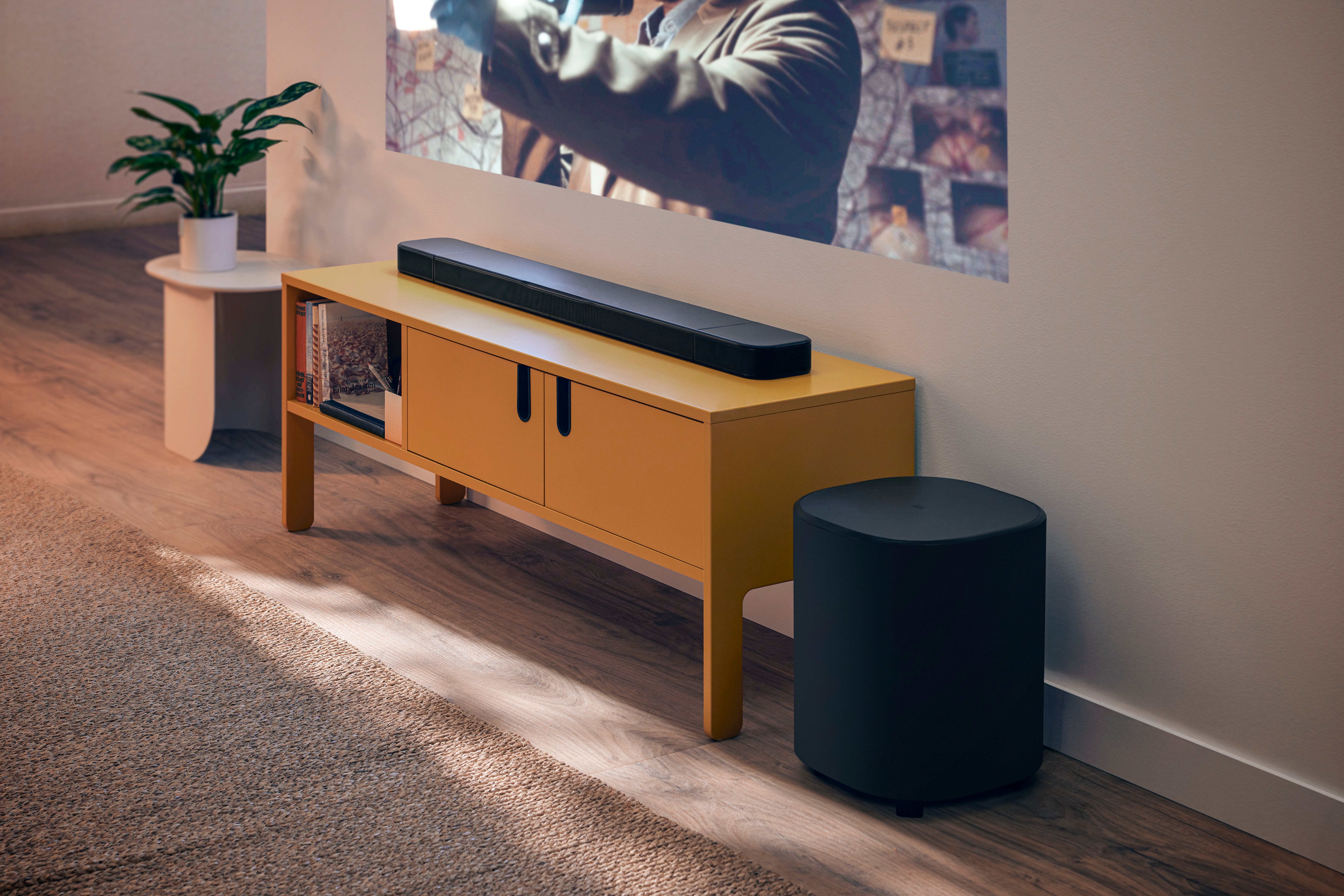 Ein modernes Wohnzimmer mit einem senffarbenen Schrank, einer Soundbar, einem Subwoofer und einer Pflanze. Ein Film wird an die Wand projiziert.