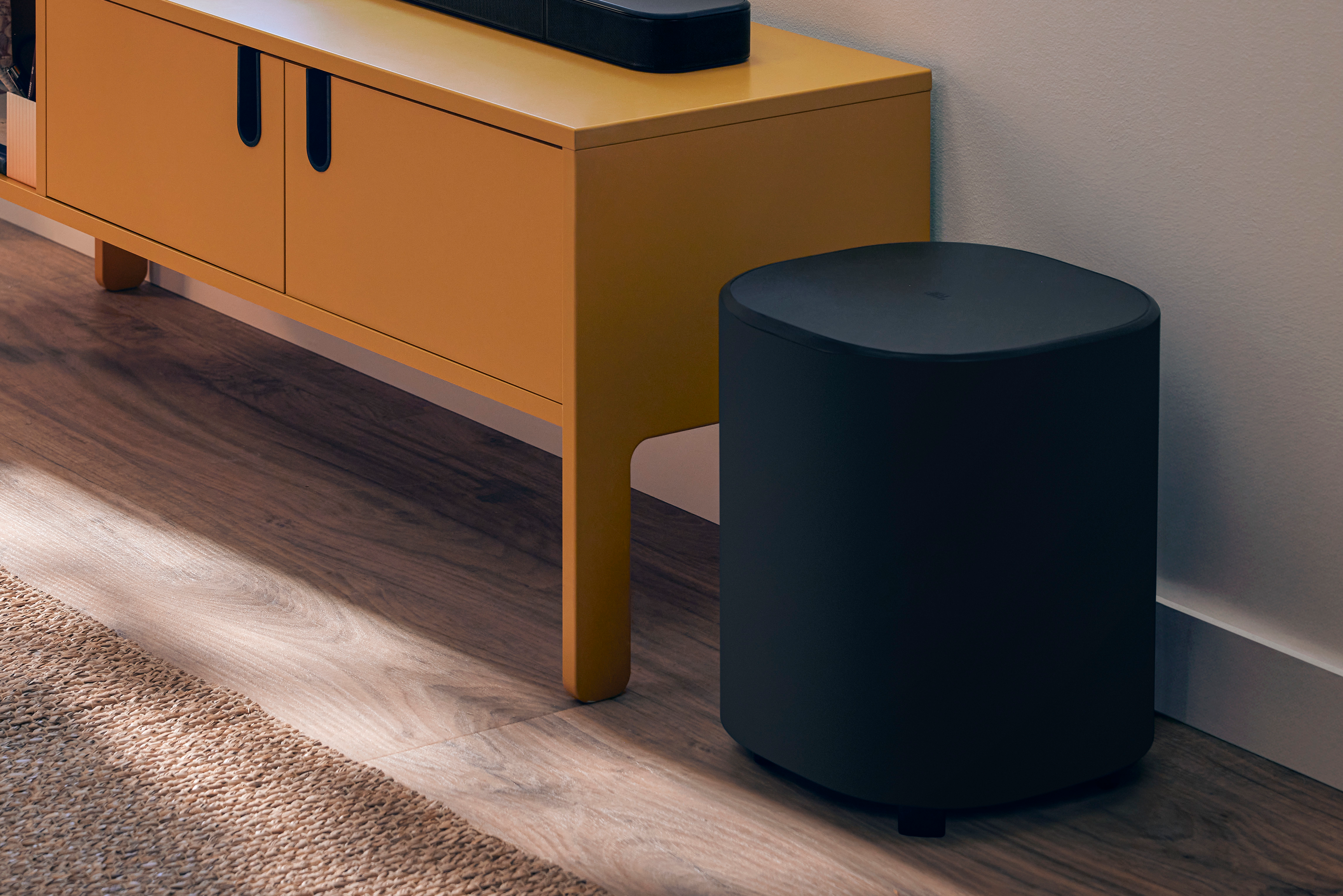 Ein schwarzer Subwoofer und ein gelber Schrank mit einer Soundbar stehen auf einem Holzboden neben einer Wand.