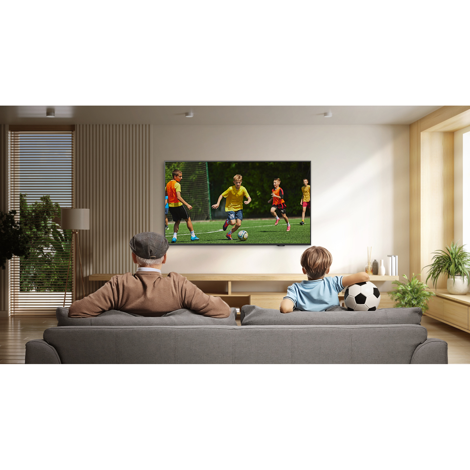 Un homme âgé et un enfant regardent un match de football à la télé. Le garçon tient un ballon de football. La pièce a des accents en bois et une plante.