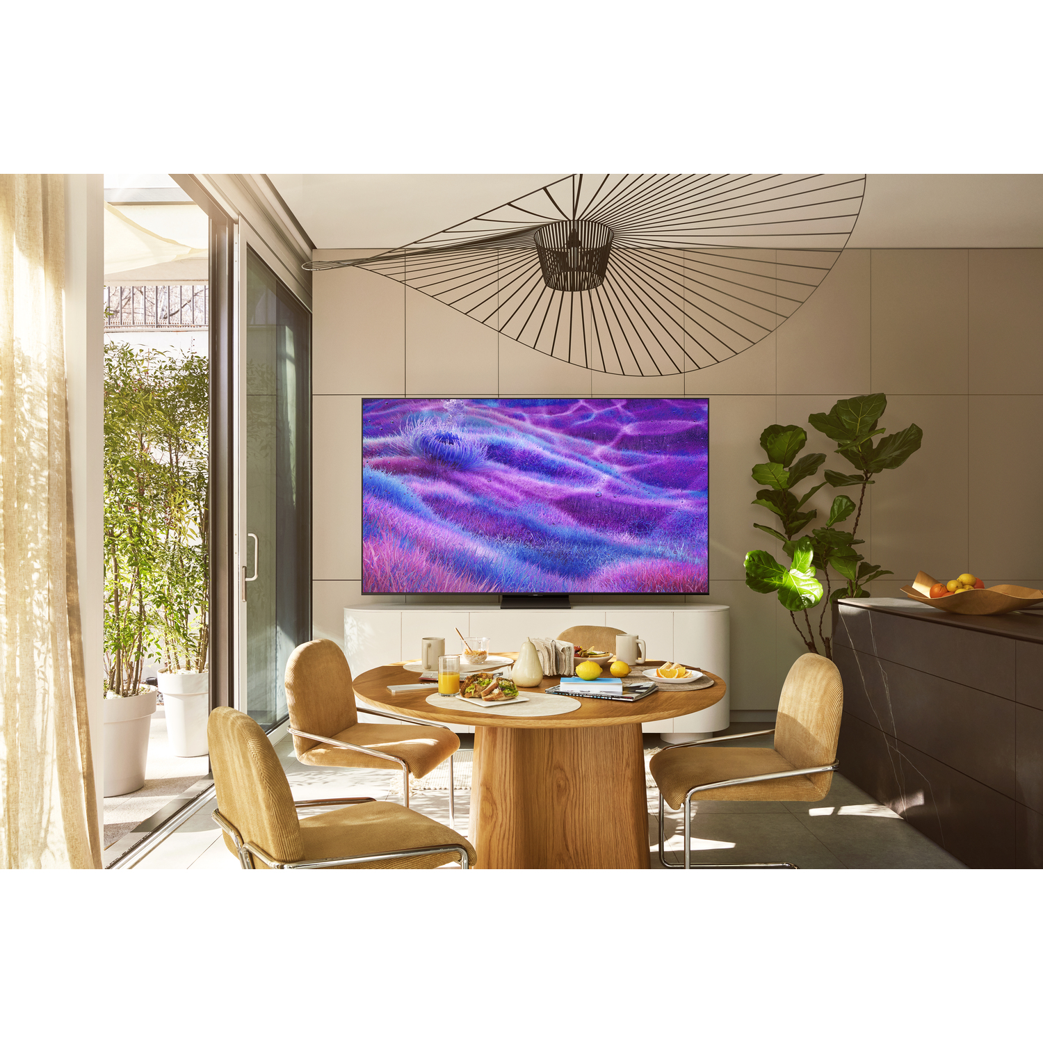 Een moderne eethoek met een ronde houten tafel gedekt voor het ontbijt. Een grote tv toont een kleurrijke abstracte afbeelding.