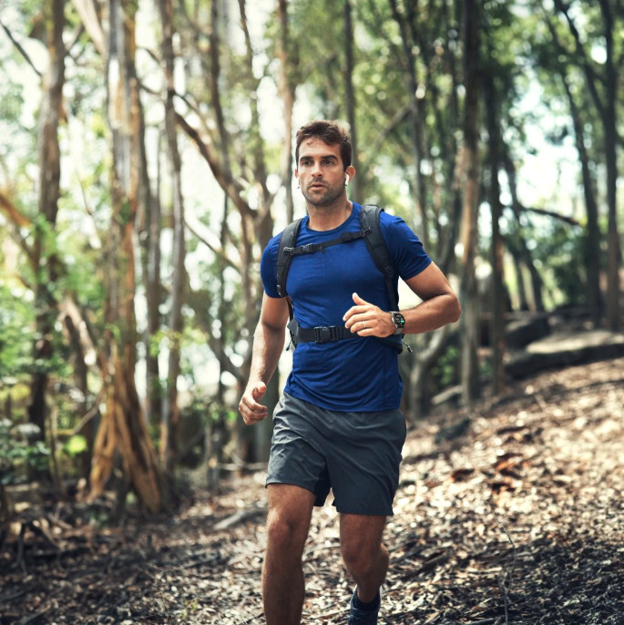 Mężczyzna biegnie ścieżką w lesie. Ma na sobie niebieską koszulkę, czarne szorty i plecak. Otaczają go drzewa.