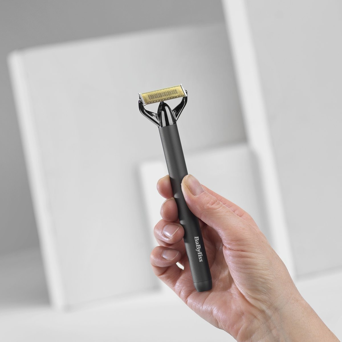 A hand holds a black and gold BaByliss razor. The background has white geometric shapes.
