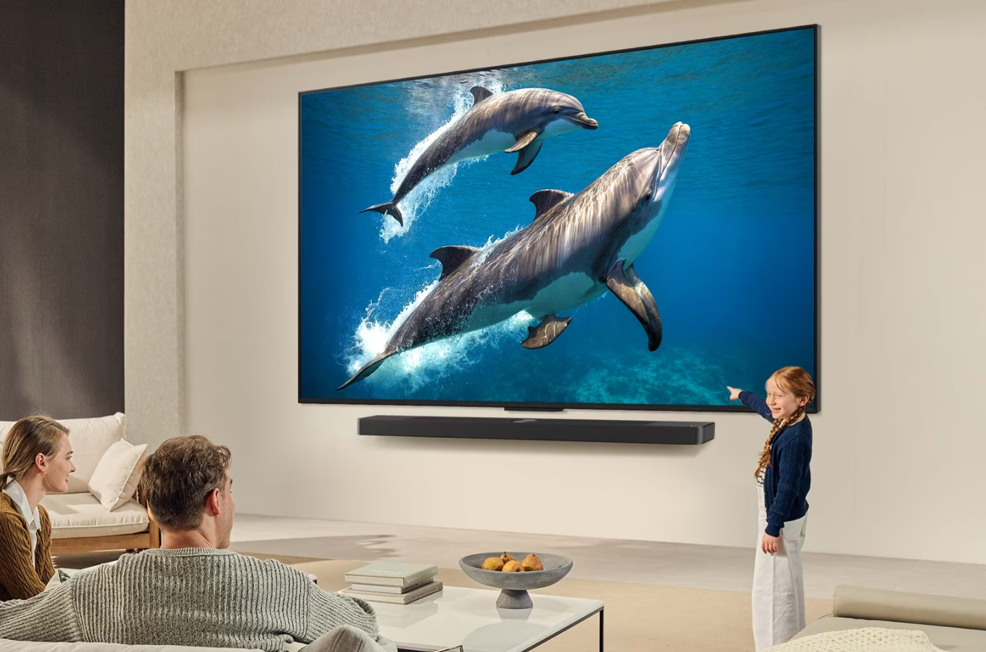 Bir aile, okyanusta yüzen yunusları gösteren bir TV izliyor. Bir kız ekranı işaret ediyor.