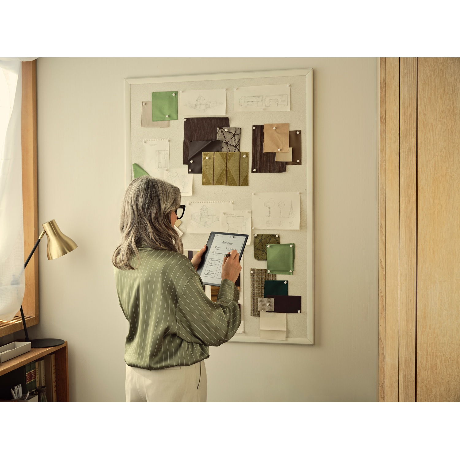 Una donna con occhiali e capelli grigi, in camicia verde, usa un tablet davanti a una mood board con campioni di tessuto e schizzi.