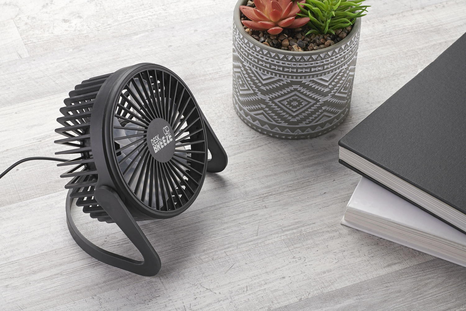 Un ventilador de escritorio negro, una suculenta en maceta y dos libros sobre una superficie de vetas de madera clara.