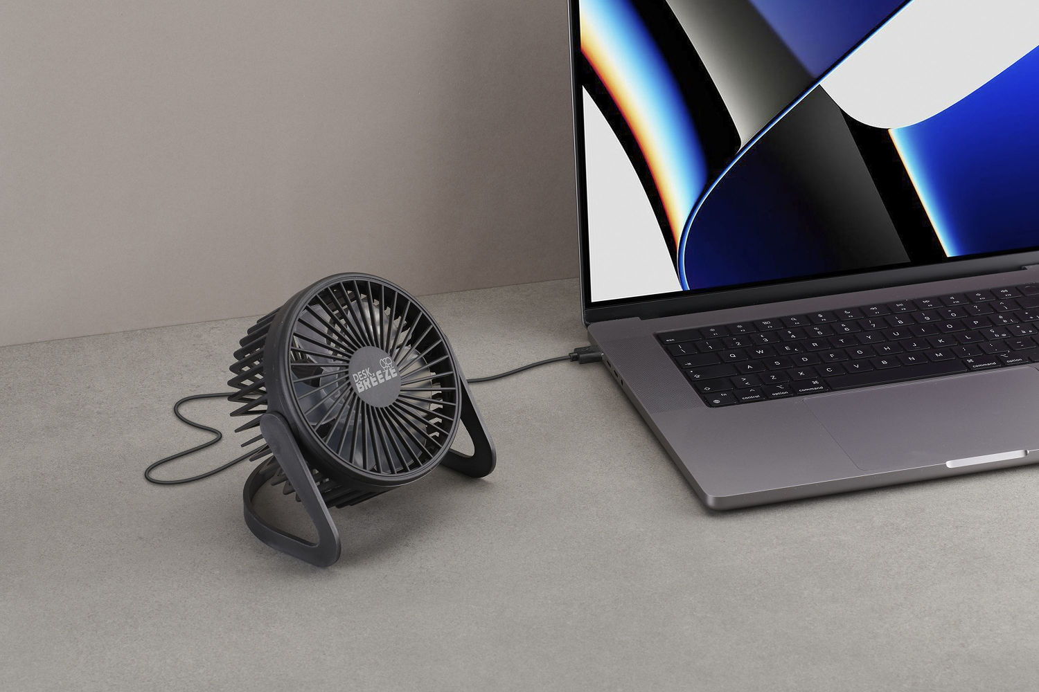 Un ventilador de escritorio negro está junto a una computadora portátil sobre una superficie gris. La computadora portátil está abierta y el ventilador está enchufado.