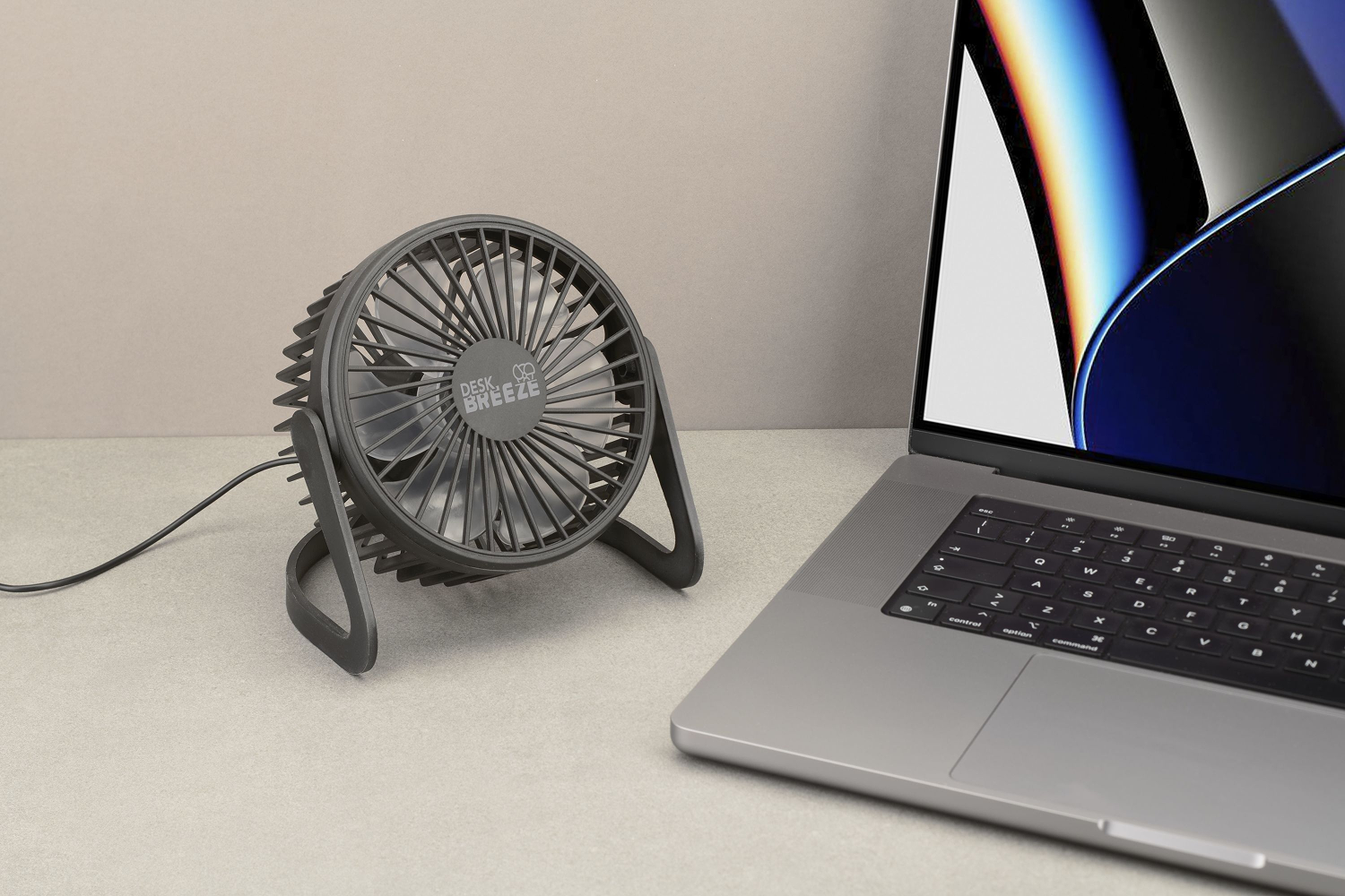Un pequeño ventilador de escritorio gris está junto a una computadora portátil sobre una superficie gris claro. El ventilador está etiquetado como "Desk Breeze".