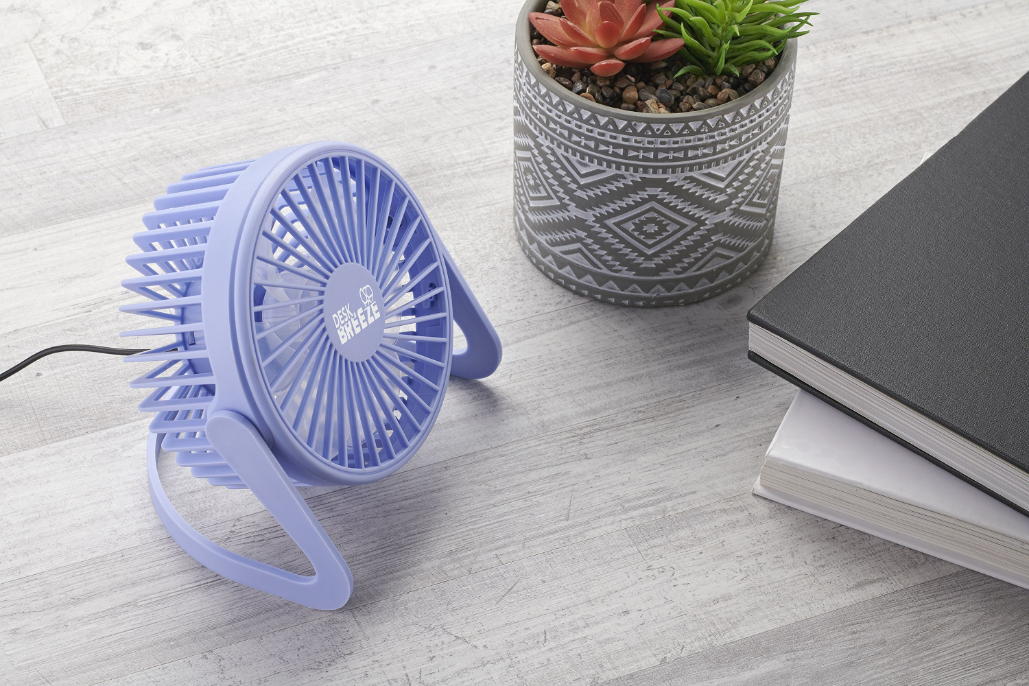 Un ventilador de escritorio morado claro, una maceta estampada con plantas y una pila de libros sobre una superficie de madera clara.