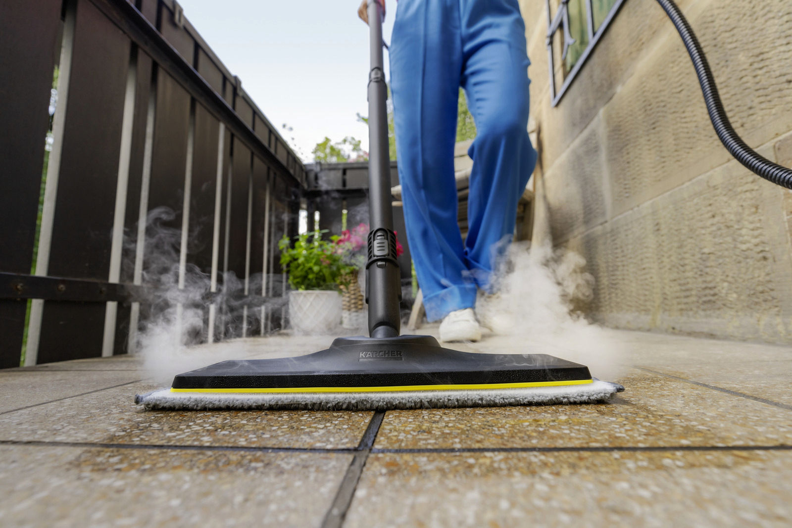 Egy kék nadrágos személy gőztisztítót használ egy csempézett teraszon. A gőz felszáll, és a tisztítón a 'KARCHER' felirat látható.