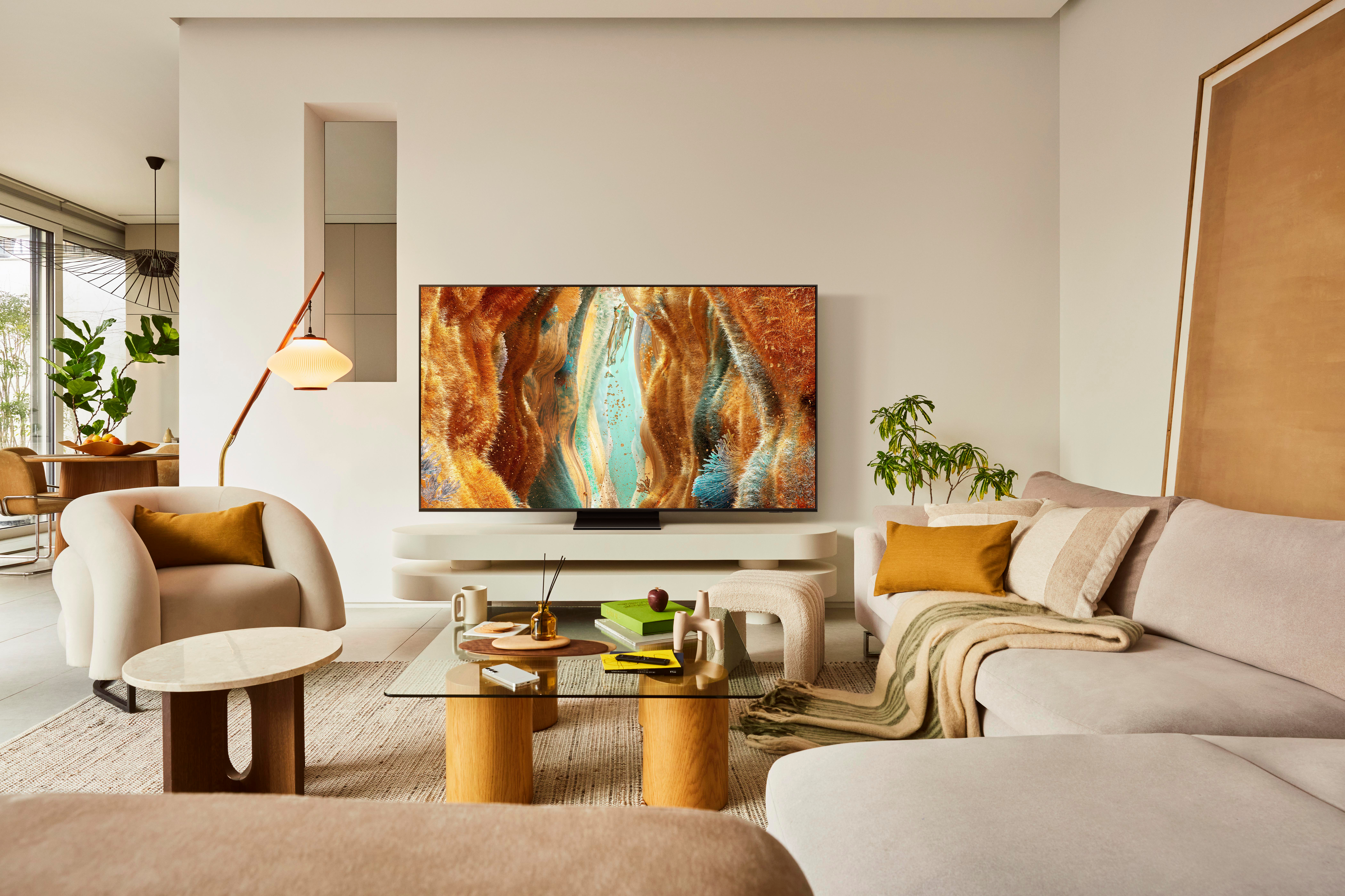 Wohnzimmer mit großem Fernseher, beigem Sofa, weißem Stuhl, Couchtisch und Pflanzen. Der Fernseher zeigt abstrakte Kunst.