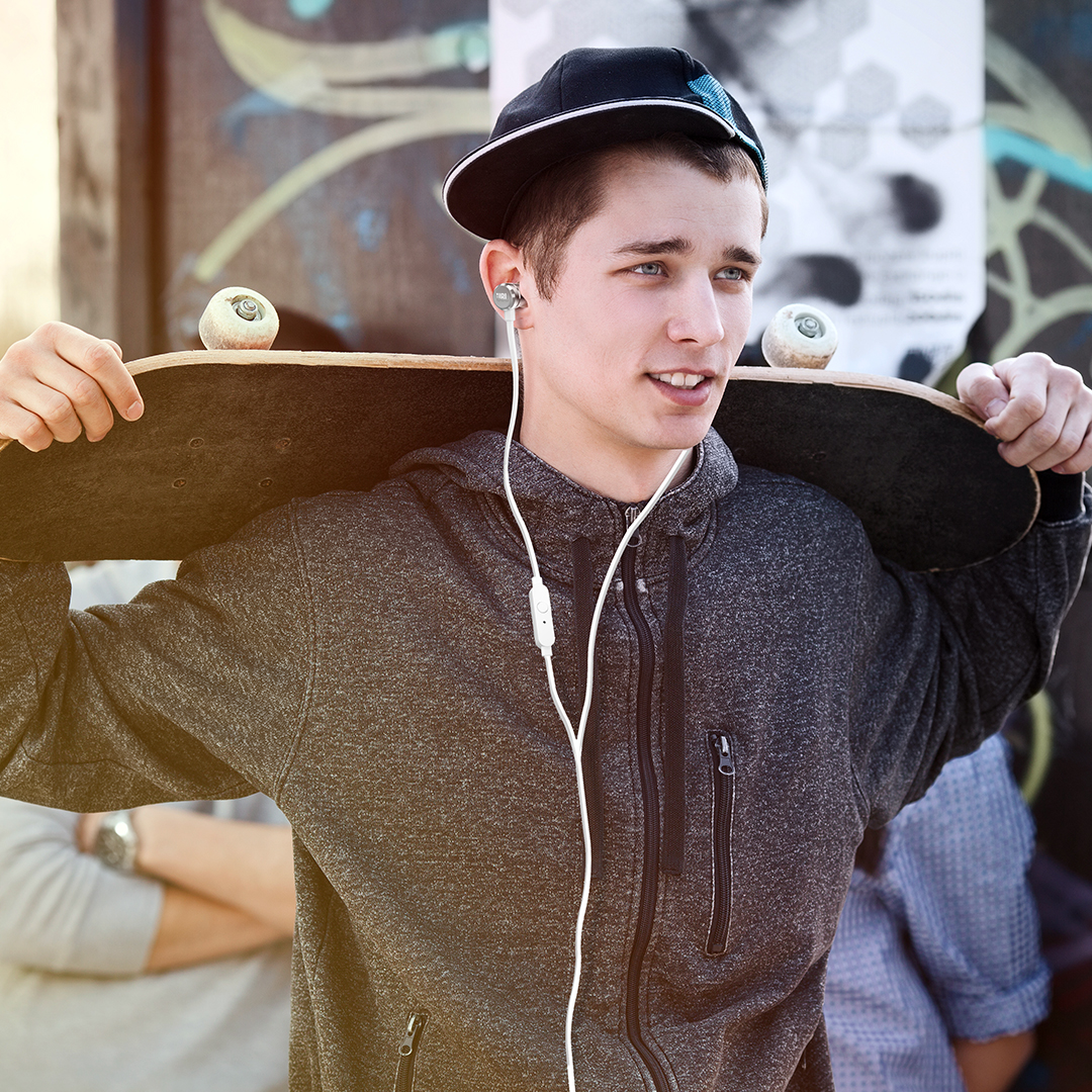 Ein junger Mann mit Mütze und Kopfhörern hält ein Skateboard. Er schaut zur Seite. Andere sind im Hintergrund.