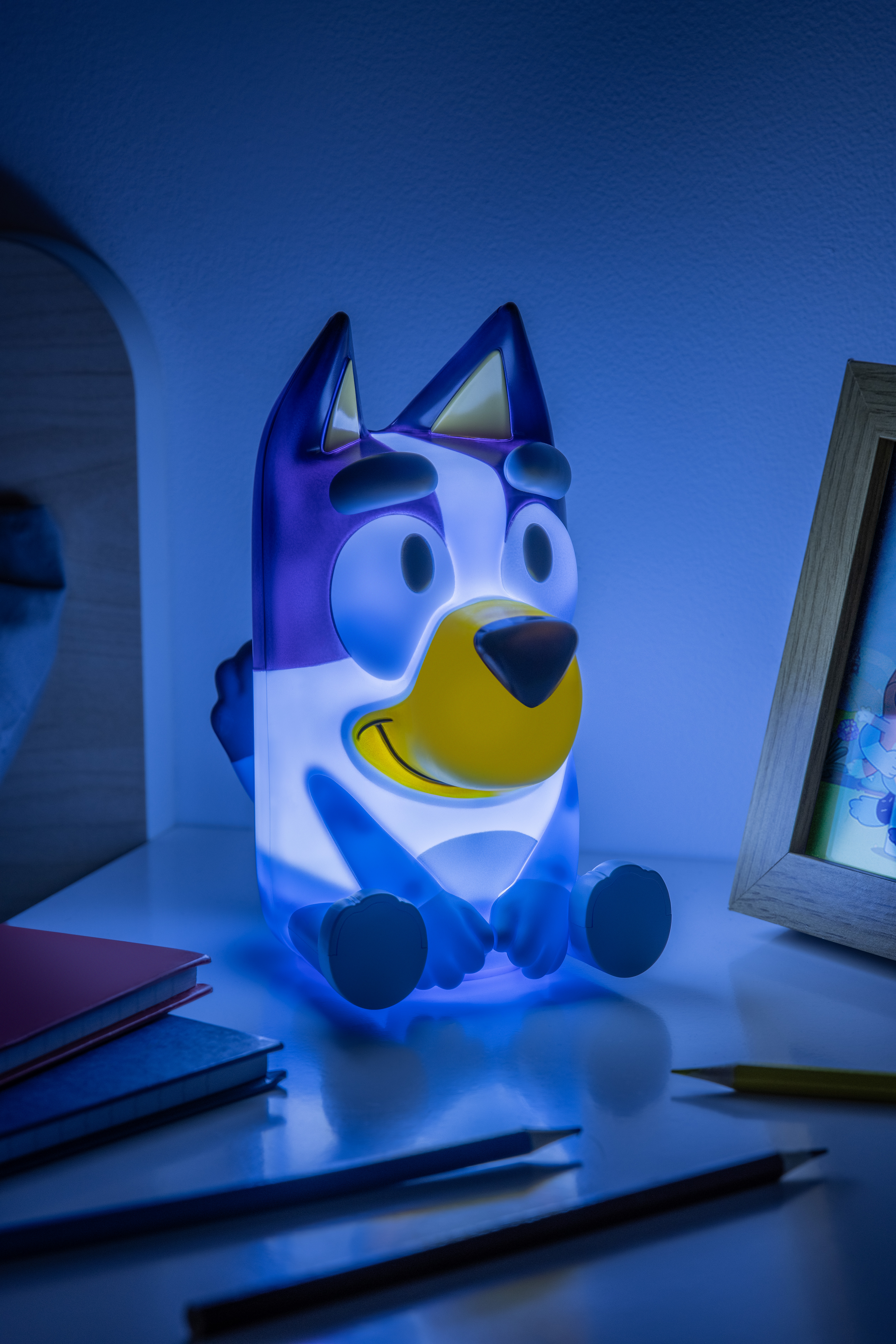 Ein Bluey-Nachtlicht steht auf einer weißen Oberfläche. Bücher und Bleistifte liegen auf der Oberfläche, und ein gerahmtes Bild ist im Hintergrund.