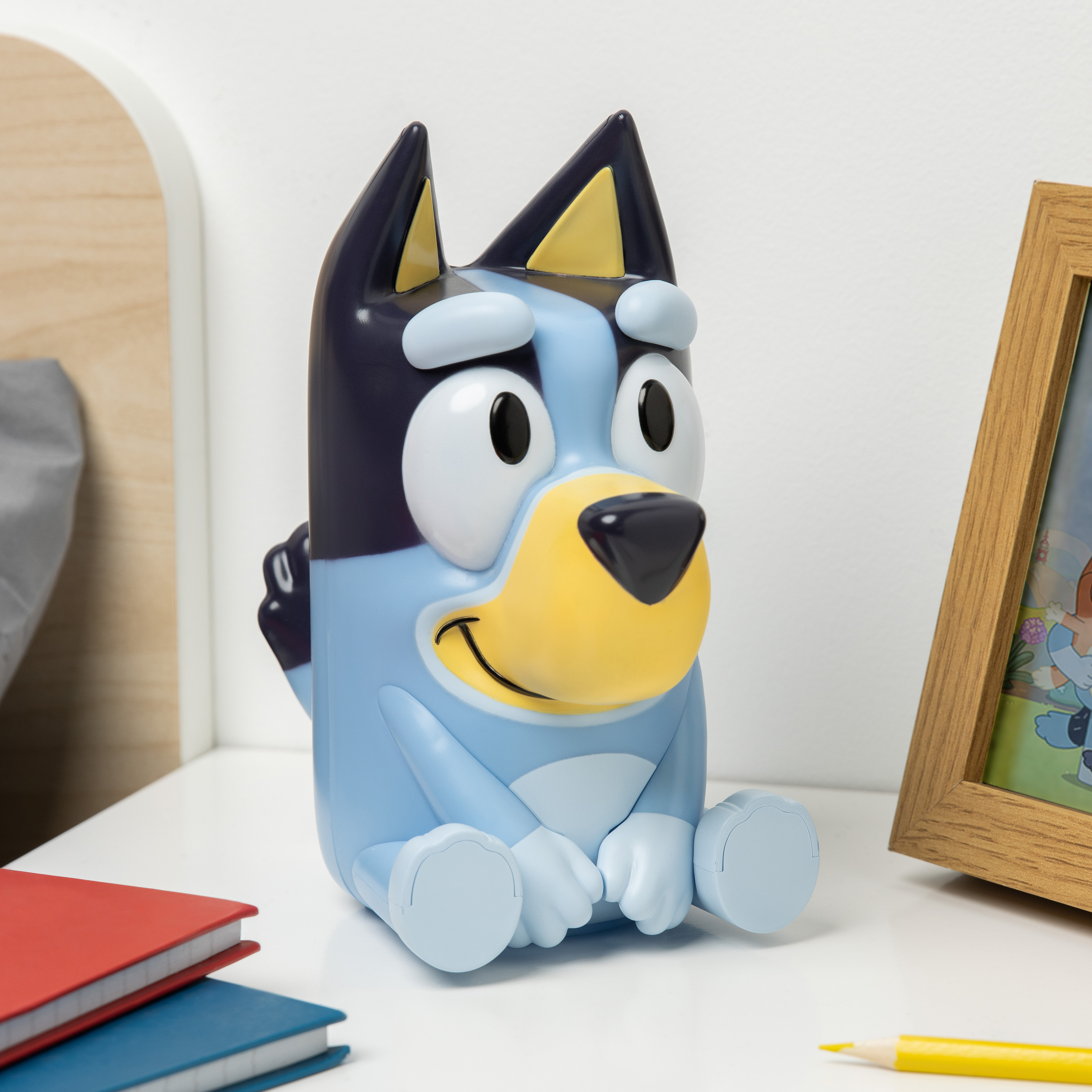 Eine Bluey-Figur sitzt auf einer weißen Oberfläche. Ein Holzrahmen mit einem Bild, Bücher und ein gelber Bleistift sind ebenfalls vorhanden.