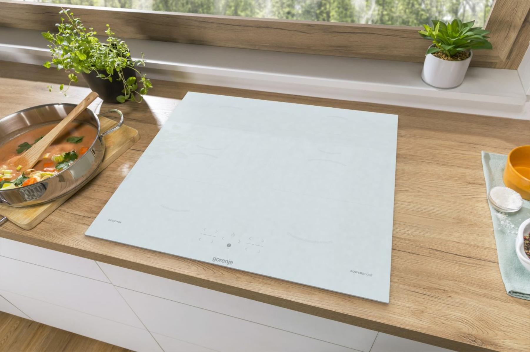 Scena kuchenna z białą płytą indukcyjną, zupą i roślinami. Drewniany blat i okno w tle.