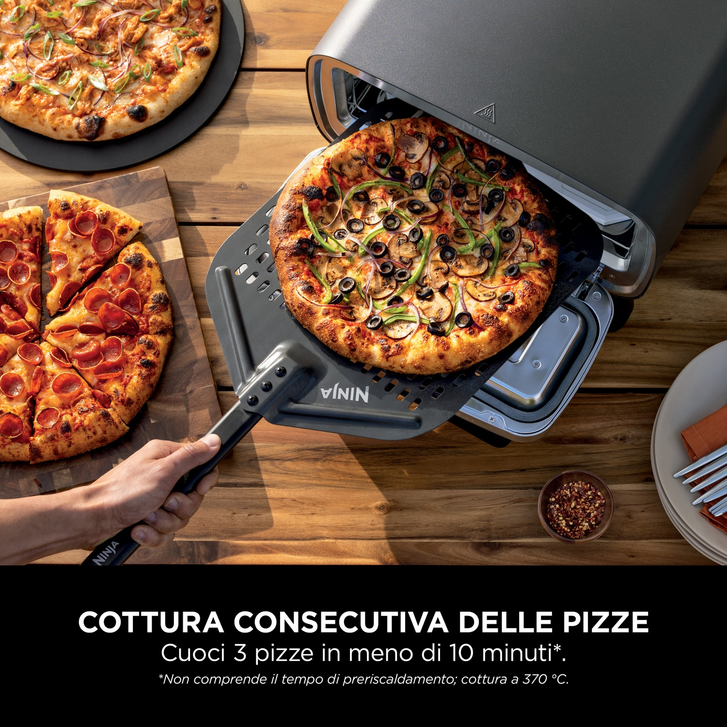 Un forno per pizza con una pizza che viene rimossa. Due pizze su un tavolo di legno sono pronte per essere mangiate. Una mano tiene la pala.
