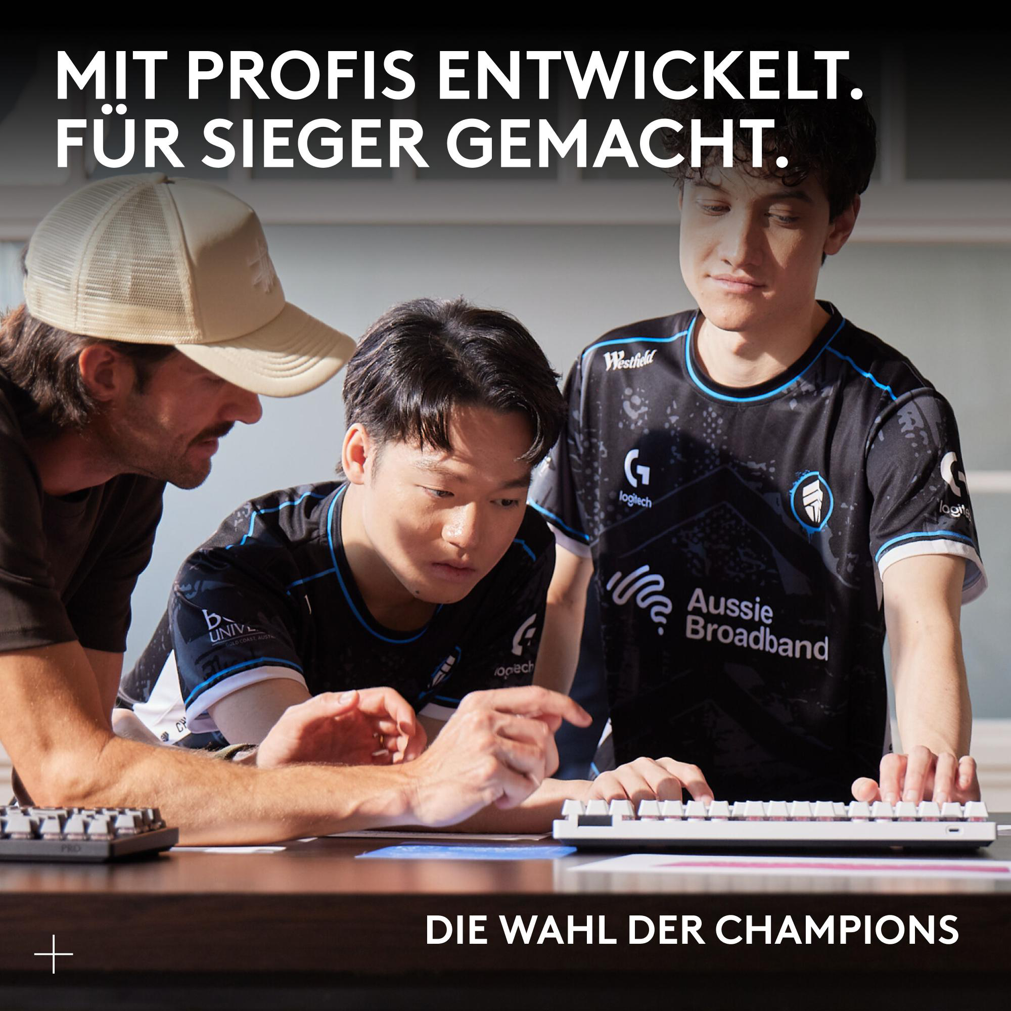Drei Personen betrachten eine Tastatur. Einer trägt eine Kappe, zwei tragen Trikots mit Logos. Der Text lautet 'Die Wahl der Champions'.