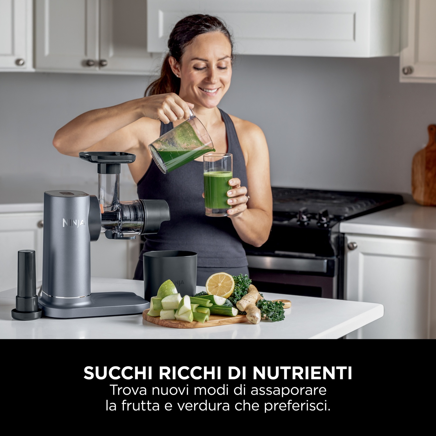 Donna che versa succo verde da una brocca in un bicchiere. L'estrattore di succo è sul bancone con frutta e verdura. Sfondo della cucina.