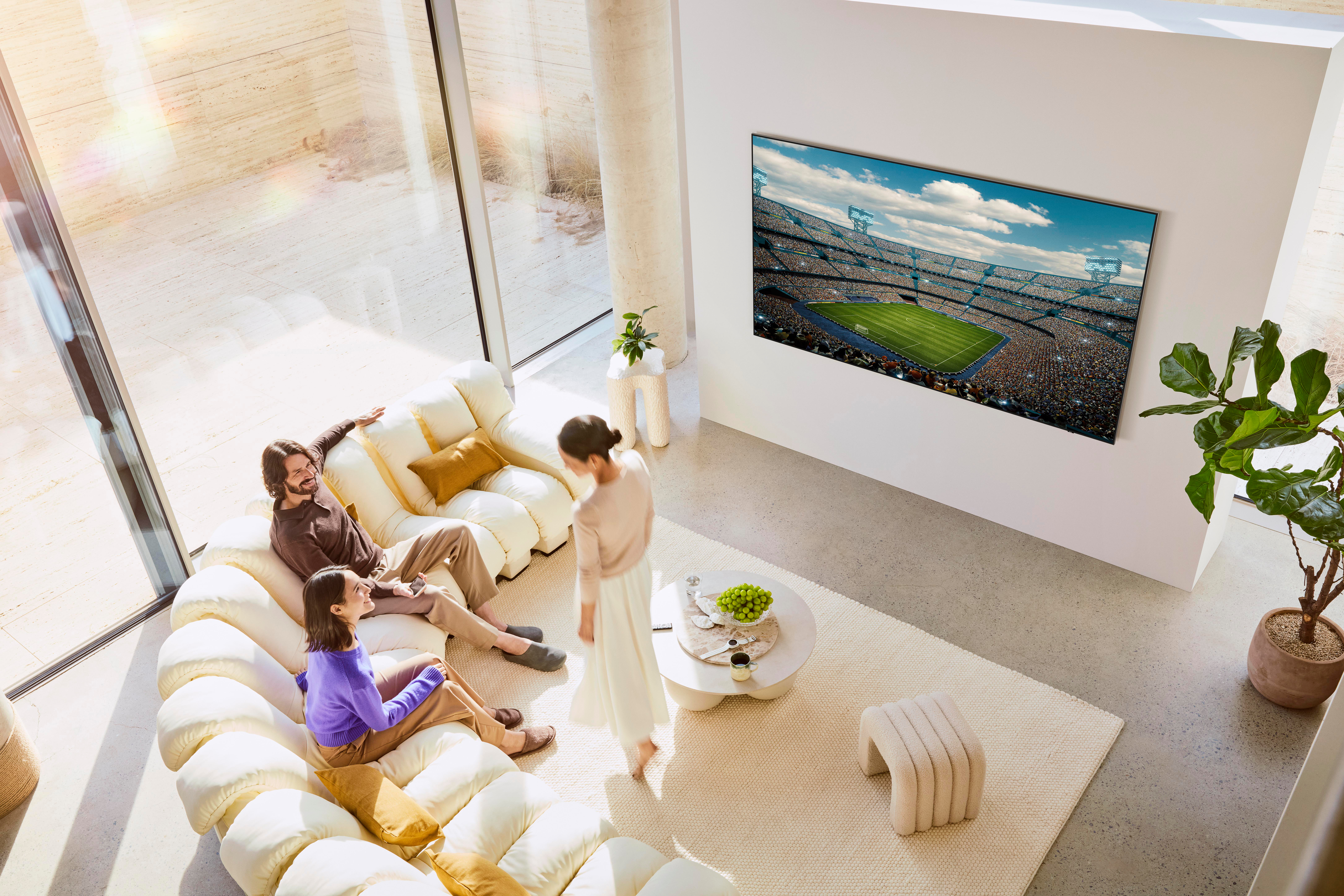 Drei Personen sehen ein Footballspiel auf einem großen Fernseher. Sie sitzen auf einem cremefarbenen Sofa. Ein weißer Teppich und eine Pflanze sind ebenfalls zu sehen.