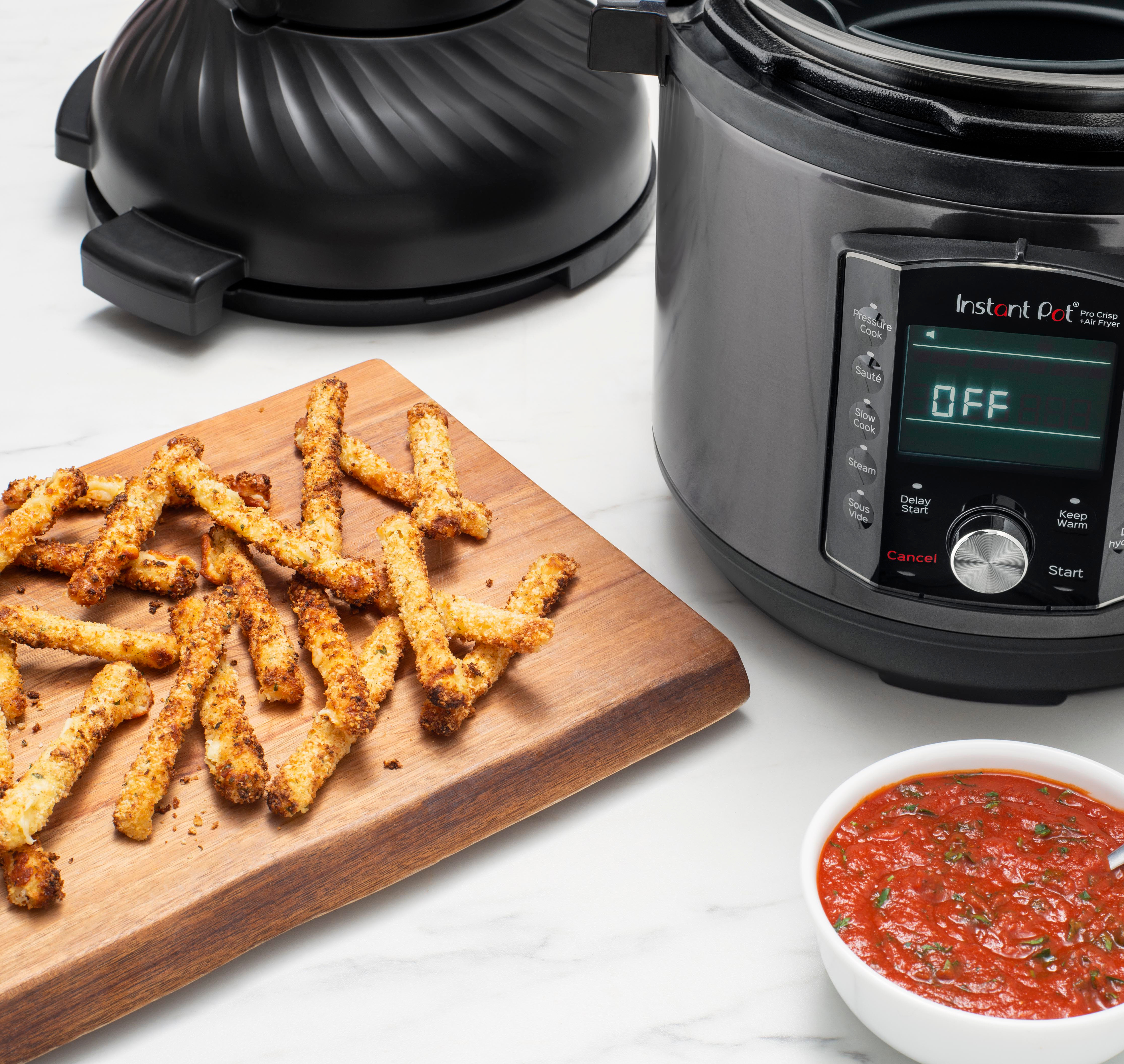 Frittierte Essensstäbchen auf einem Holzbrett, Instant Pot, Fritteuse und eine Schüssel Soße auf einem weißen Marmortisch.