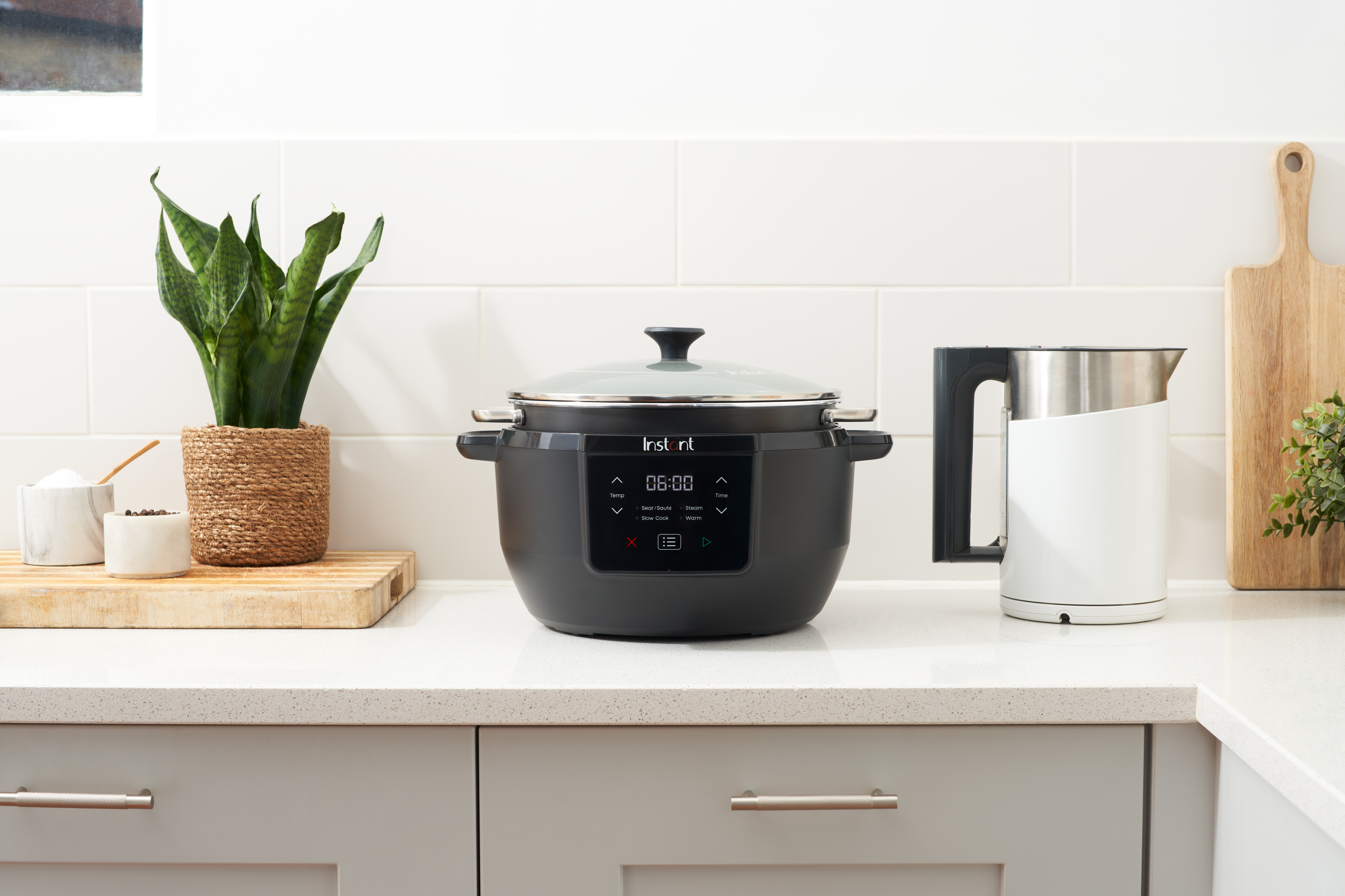 Küchenszene mit einem schwarzen Instant Pot, weißem Wasserkocher, Pflanze und Schneidebrett auf einer Arbeitsplatte. Weiße Fliesen im Hintergrund.