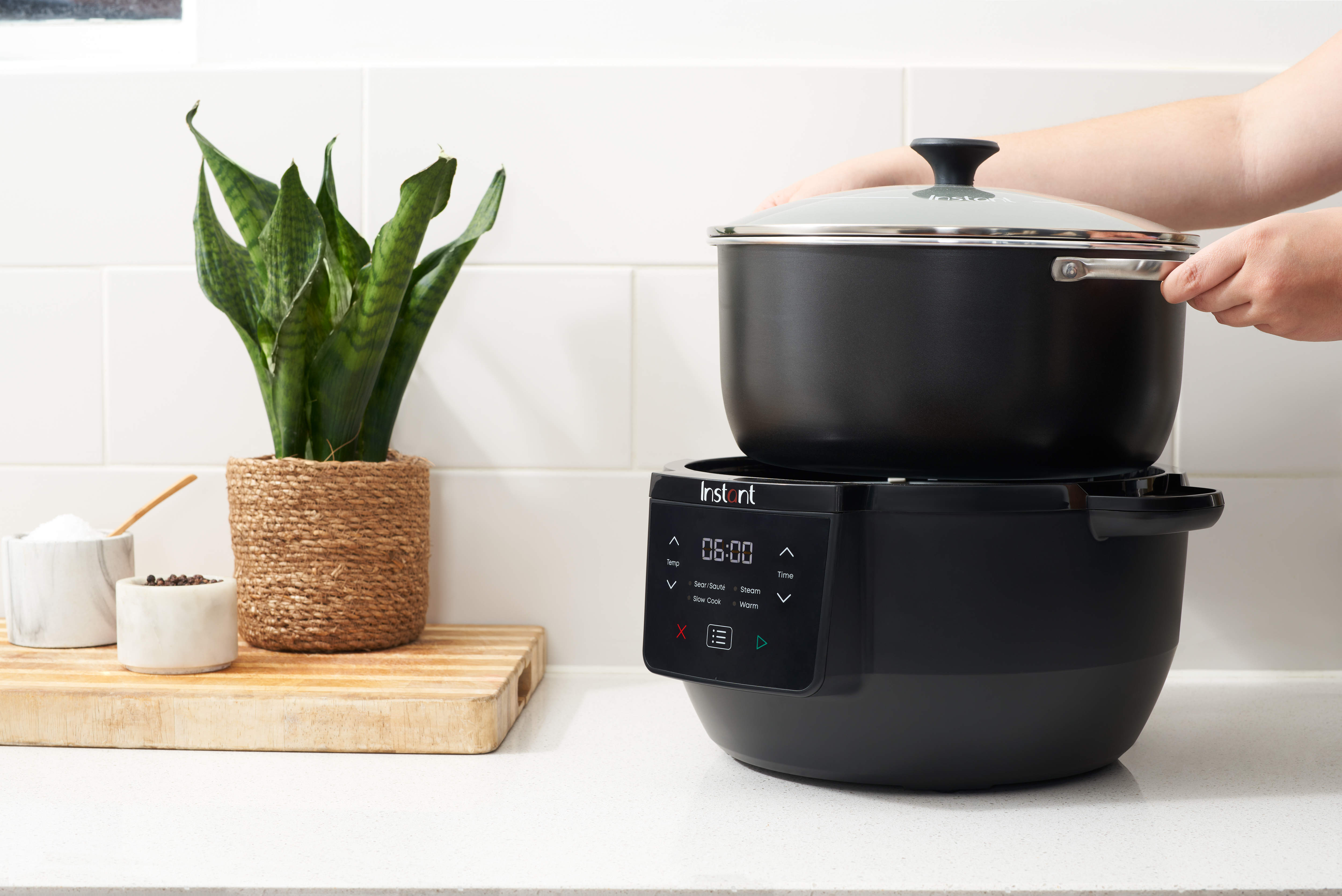 Ein schwarzer Instant Pot mit Deckel, der von einer Person gehalten wird. Links stehen eine Pflanze in einem gewebten Topf und Küchenutensilien.