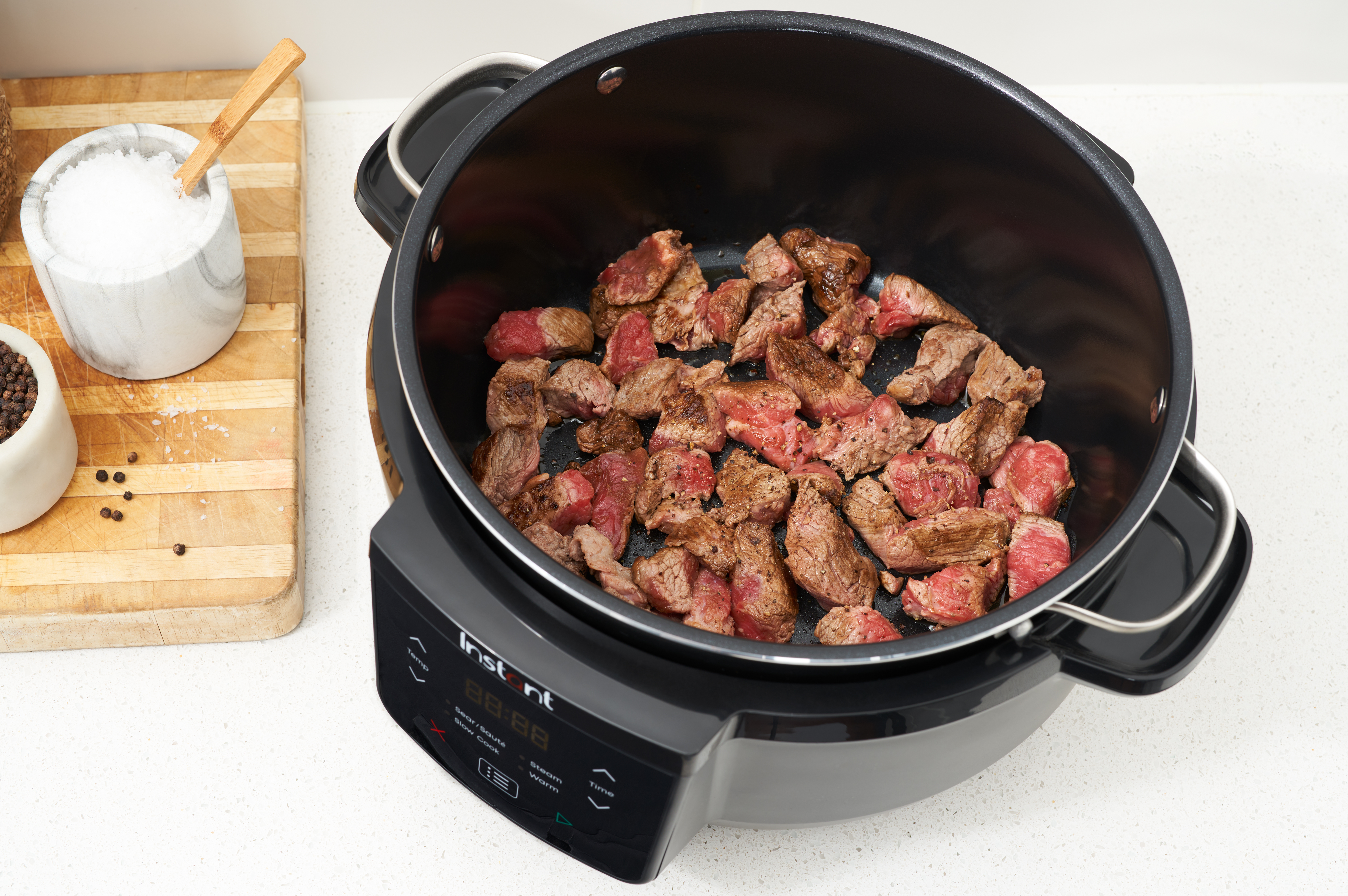 Ein schwarzer Instant Pot mit gekochten Rindfleischwürfeln. Salz und Pfeffer sind links. Ein Holzschneidebrett ist ebenfalls sichtbar.