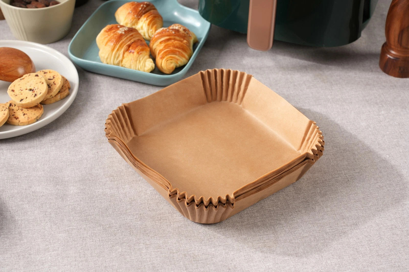 Brązowe papierowe kubeczki do pieczenia ułożone na szarej tkaninie. W tle rogaliki, ciasteczka i inne produkty spożywcze.