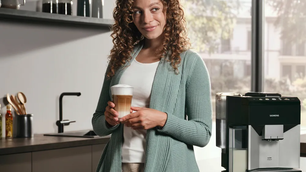 Egy nő lattét tart egy konyhában. Zöld kardigánt visel, és mosolyog. Jobbra egy kávéfőző gép látható.