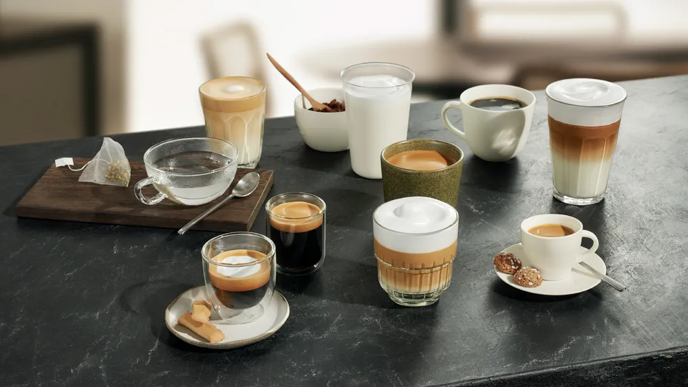 Különböző kávéitalok, tea és snackek sötét pulton elrendezve. Lattékat, eszpresszókat és süteményeket tartalmaz.