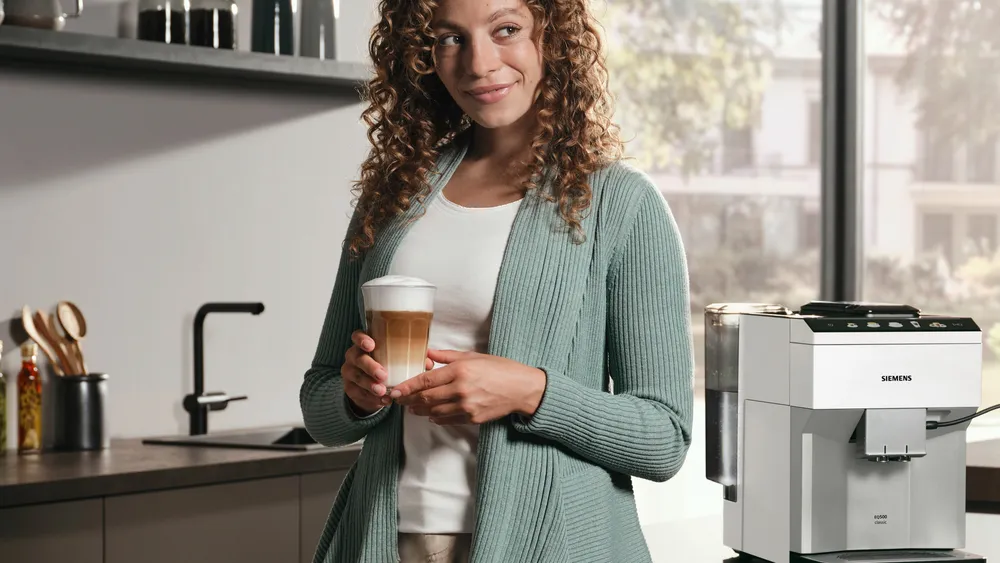 Egy nő a konyhában lattét tart. Jobbra egy kávéfőzőgép van. A háttér egy mosogatóval ellátott konyha.