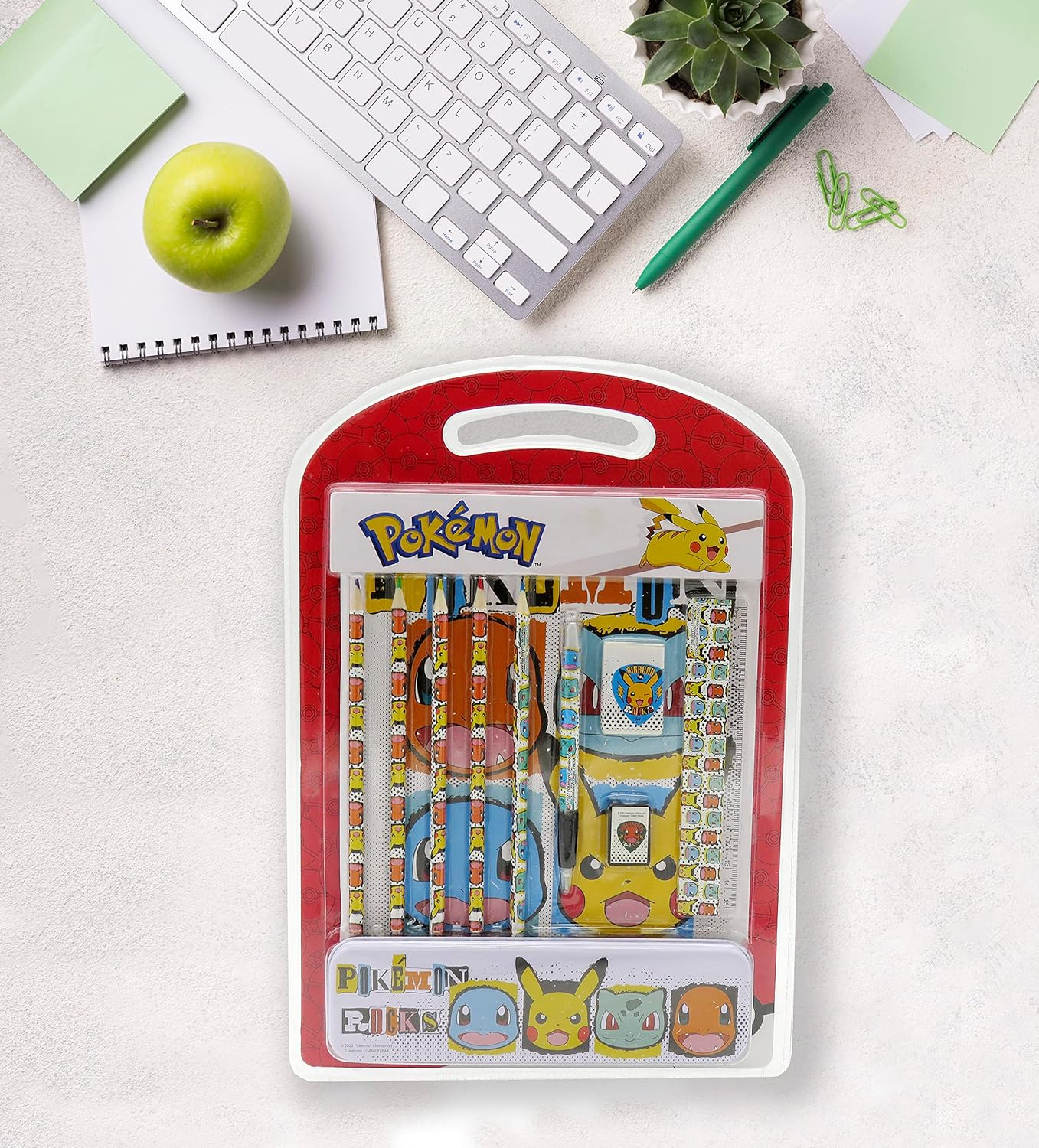 Una configuración de escritorio con un estuche de lápices con tema de Pokémon y útiles escolares, incluyendo un teclado, una manzana y un bolígrafo. Fondo blanco.