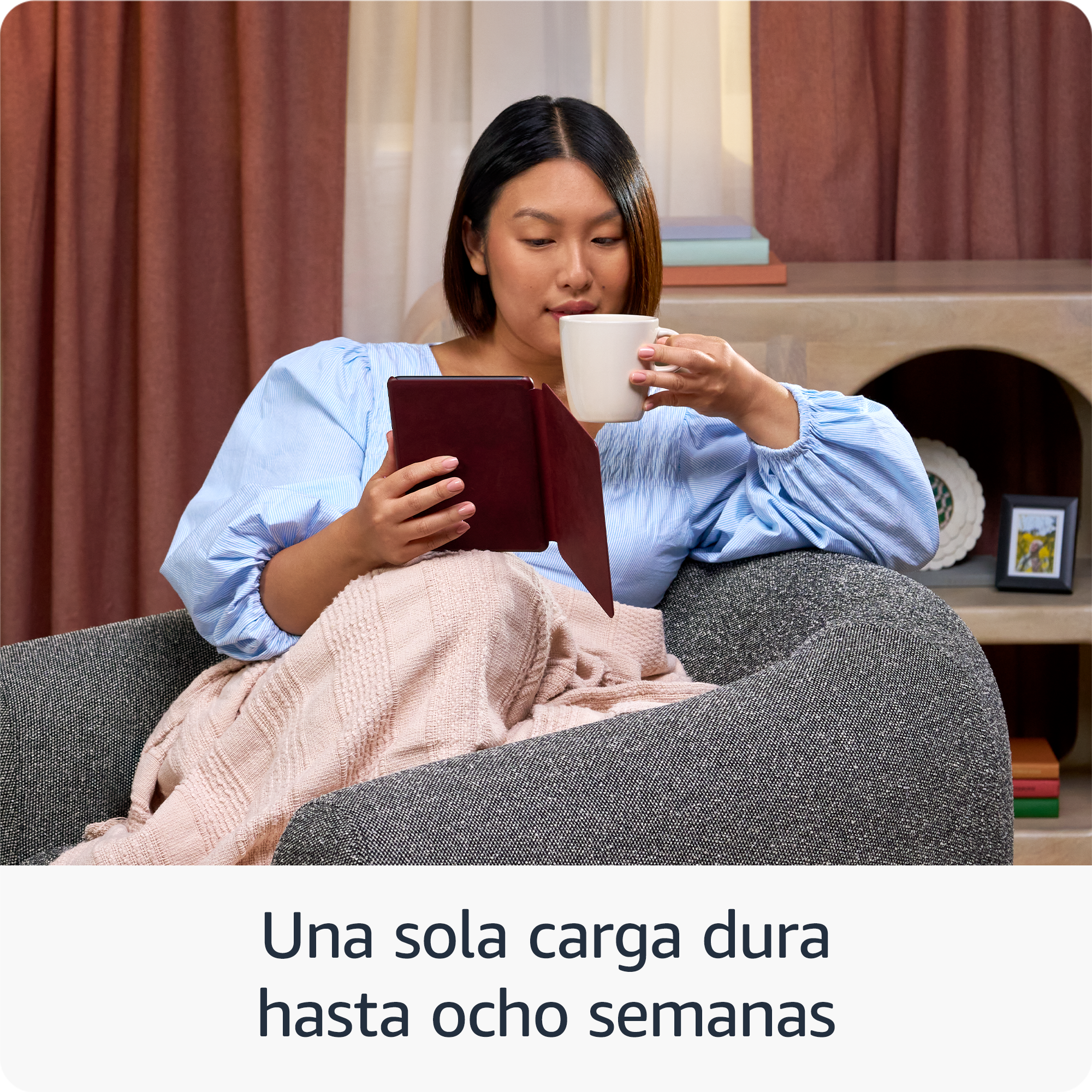 Mujer leyendo tableta, sosteniendo taza, sentada en sillón gris. Manta rosa, cortinas marrones y libros en el fondo.