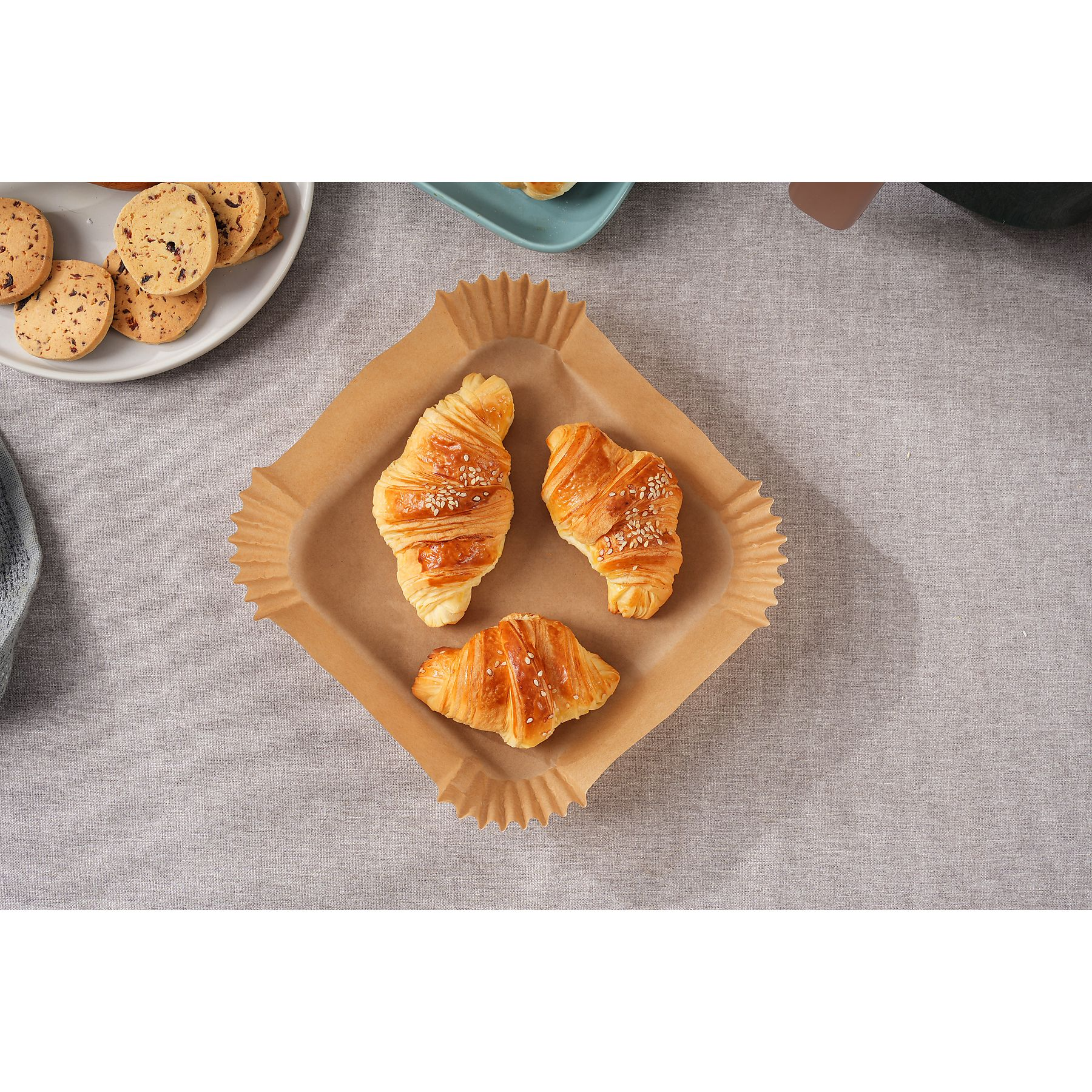 Trzy rogaliki na brązowej papierowej tacy, otoczone ciastkami na białym talerzu, na szarym obrusie.