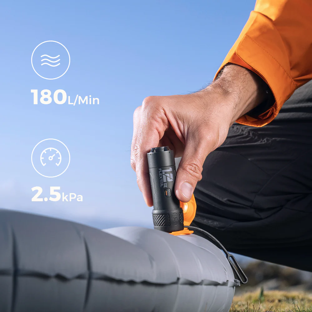 Egy személy egy szürke felfújható matracot fúj fel egy fekete pumpával. A pumpa 180L/Min és 2,5kPa értéket mutat. Kék ég a háttérben.