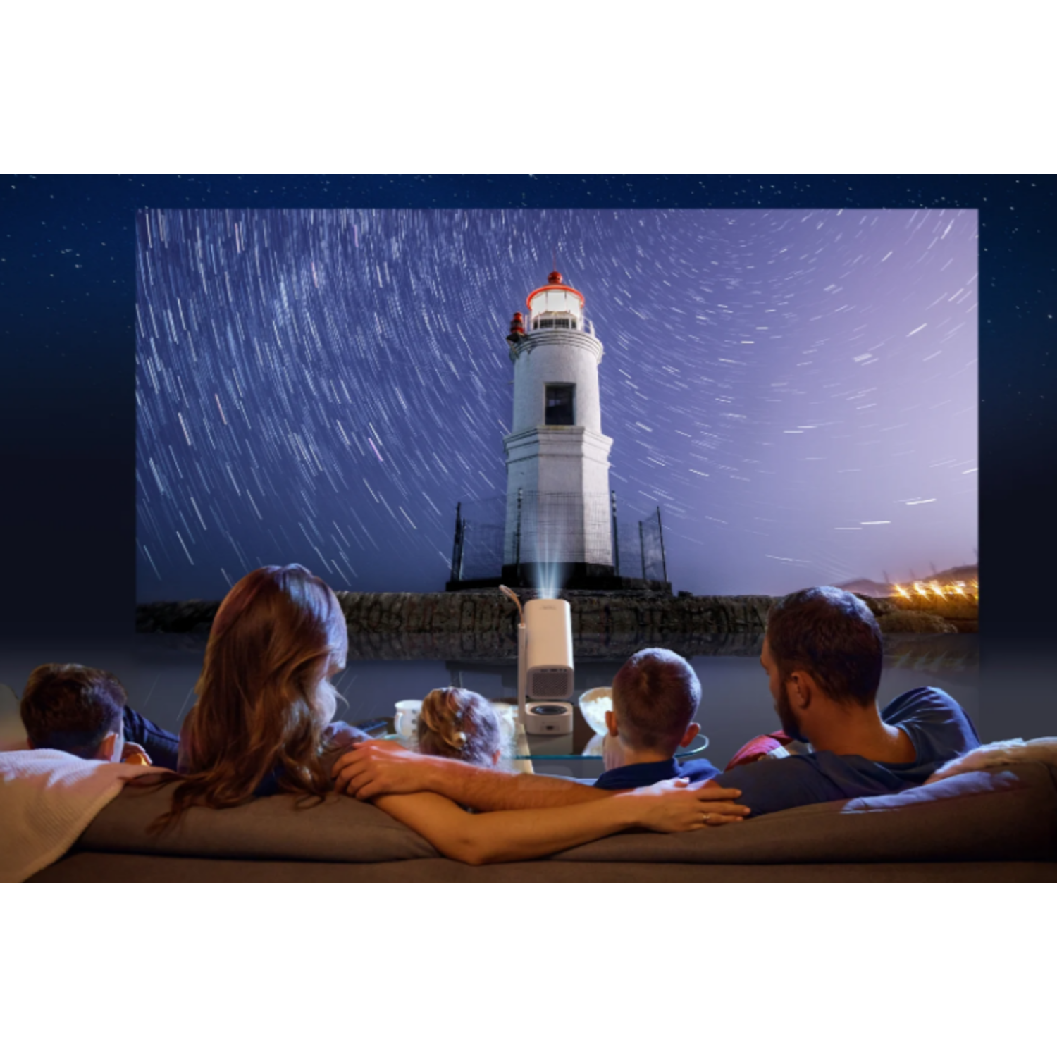 Familie sieht ein projiziertes Bild eines Leuchtturms unter Sternenhimmel. Sie sitzen auf einer Couch mit dem Projektor davor.