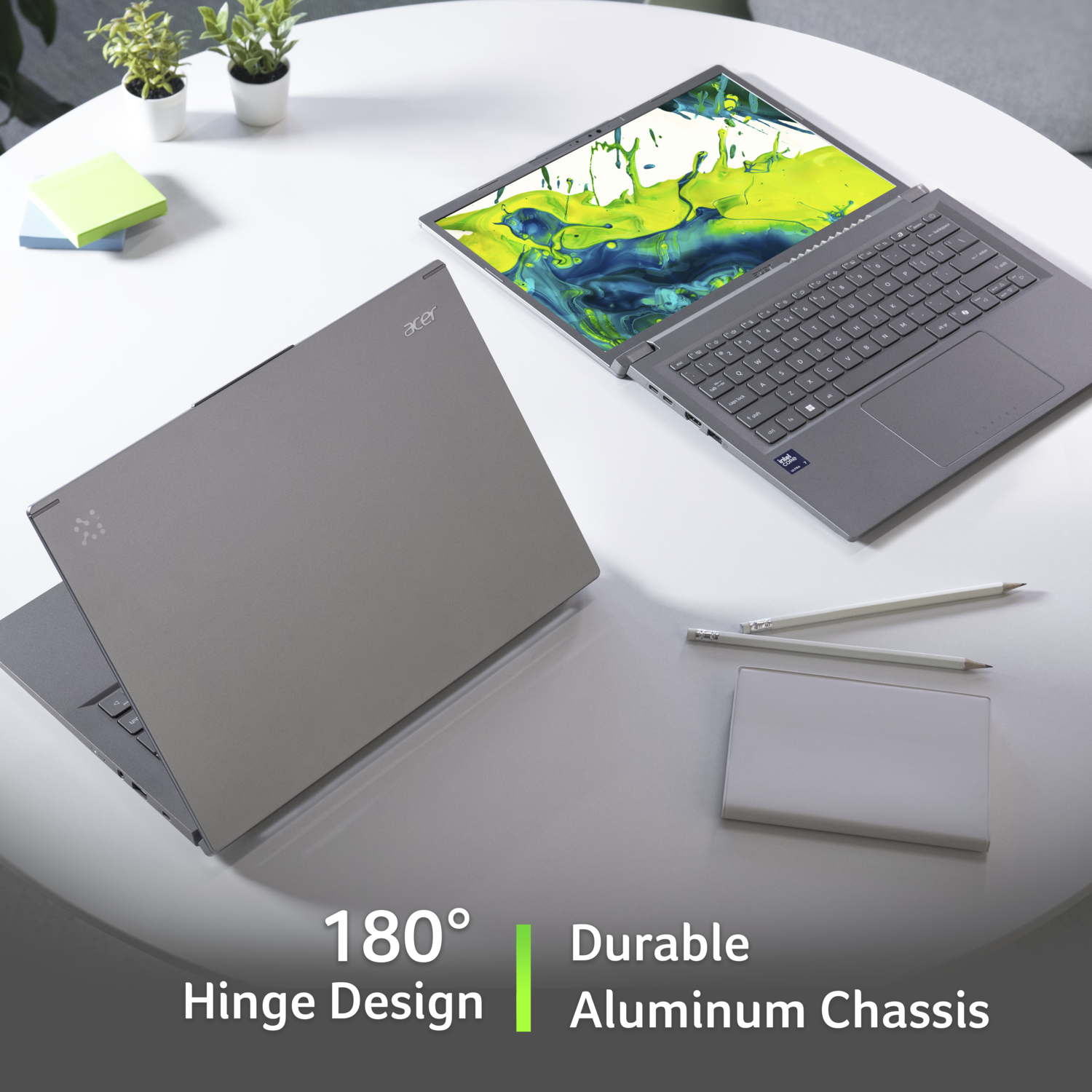 Two Acer laptops on a white table, one open. Pencils and a pad are also present. Plants are in the background.