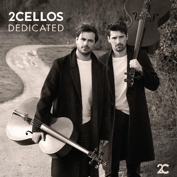 Zwei Männer in schwarzen Mänteln halten Celli. Ein Cello liegt am Boden, das andere wird aufrecht gehalten. Im Hintergrund ist ein Weg.
