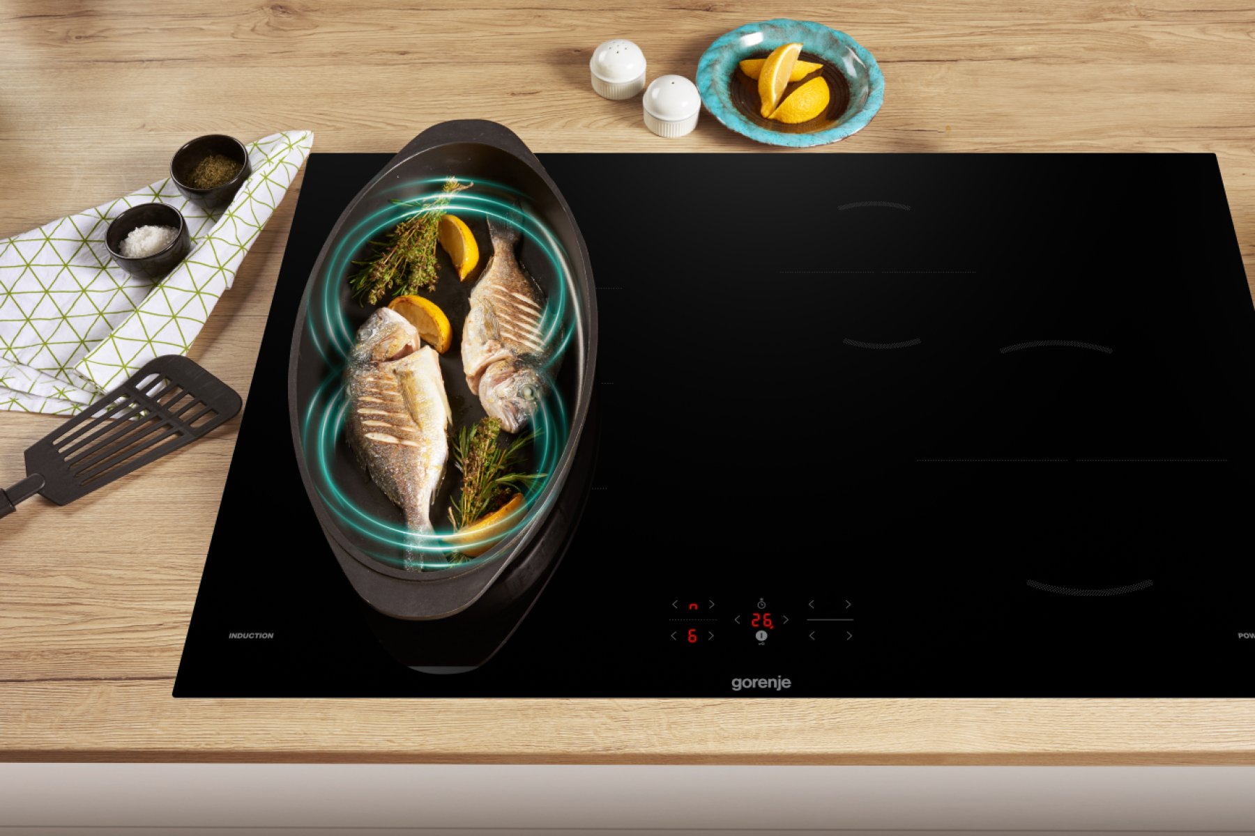 Ryba z cytryną w patelni na czarnej płycie indukcyjnej. Sól, pieprz i talerz z cytryną na drewnianej powierzchni.