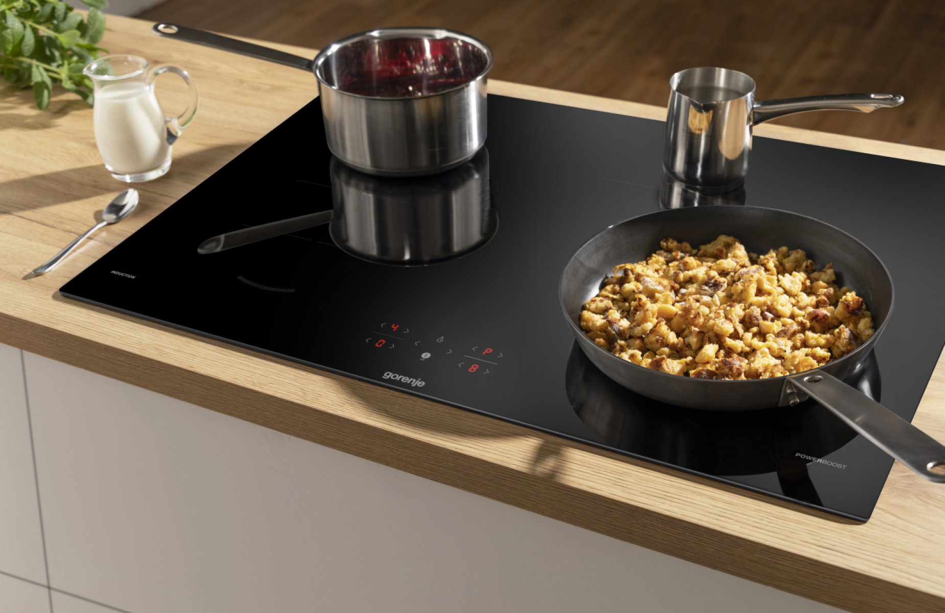 Scena kuchenna z czarną płytą indukcyjną. Na kuchence patelnie z jedzeniem. Dzbanek z mlekiem, łyżka i zioła z boku.